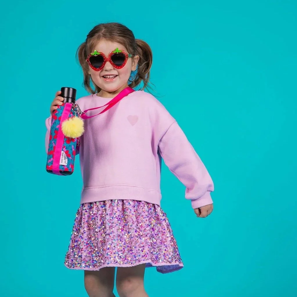 A young girl with pigtails wearing pink heart-shaped sunglasses, a light pink sweatshirt, and a sparkly purple skirt. She is smiling and holding a Bottlesoc with a colorful design and a pink strap, against a blue background.