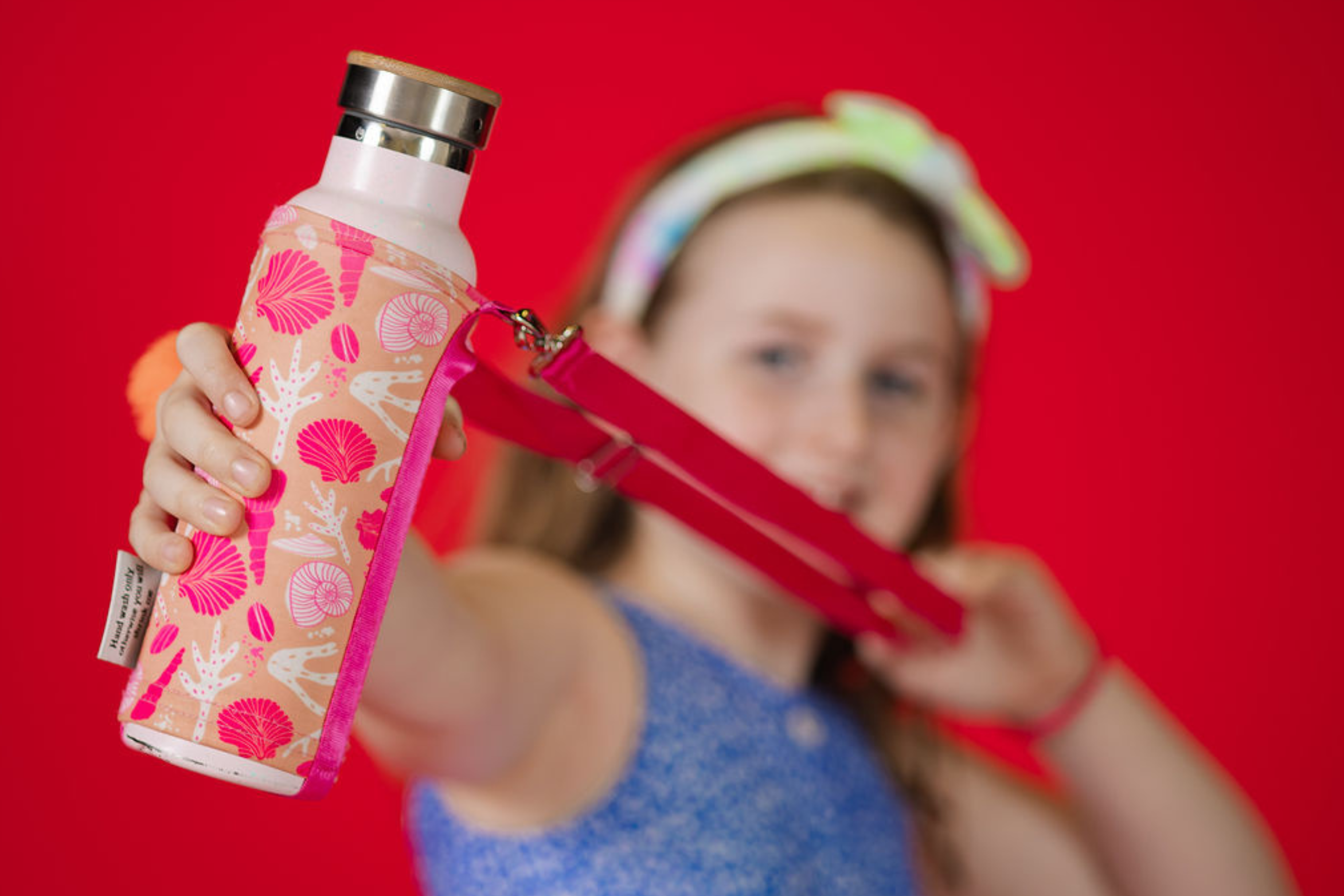 A young woman holding a pink and white water bottle with a sea shell design and a pink strap, against a red background.