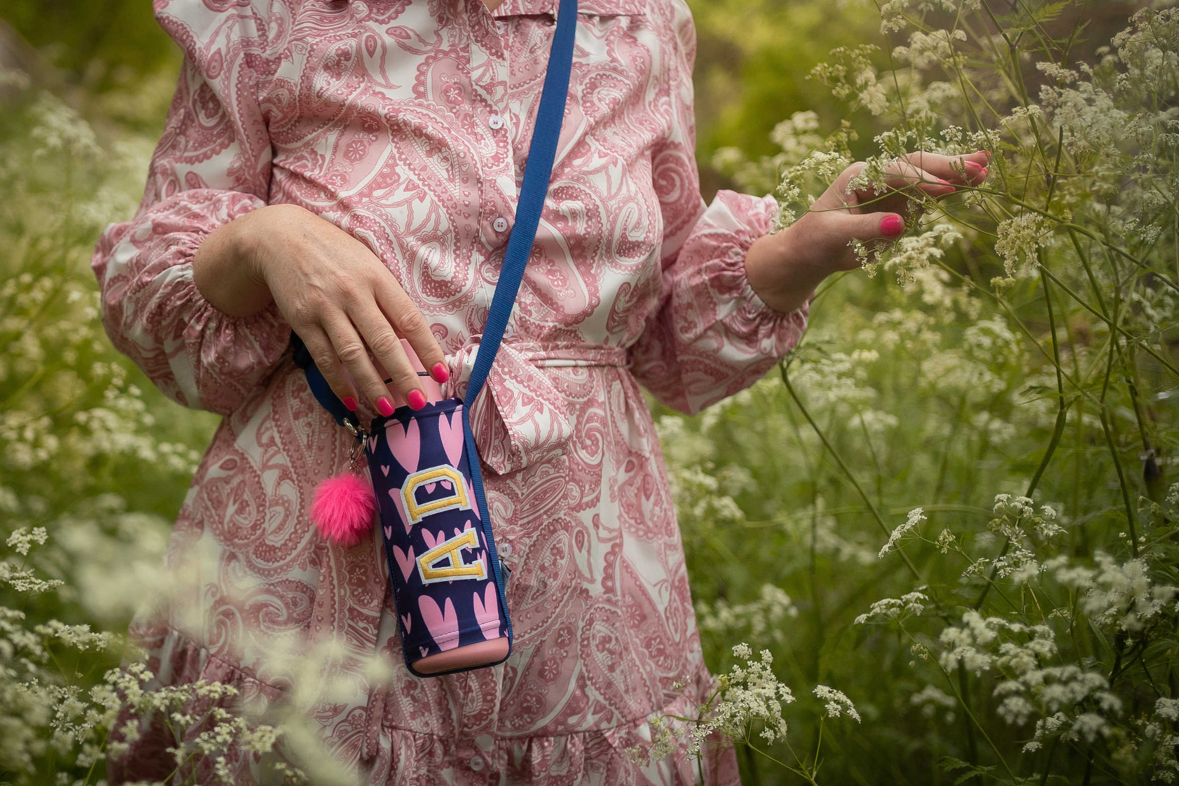 A woman in a pink patterned dress with pink nails and a blue strap bag with a pink pom-pom is standing among white flowering plants, gently touching the flowers.