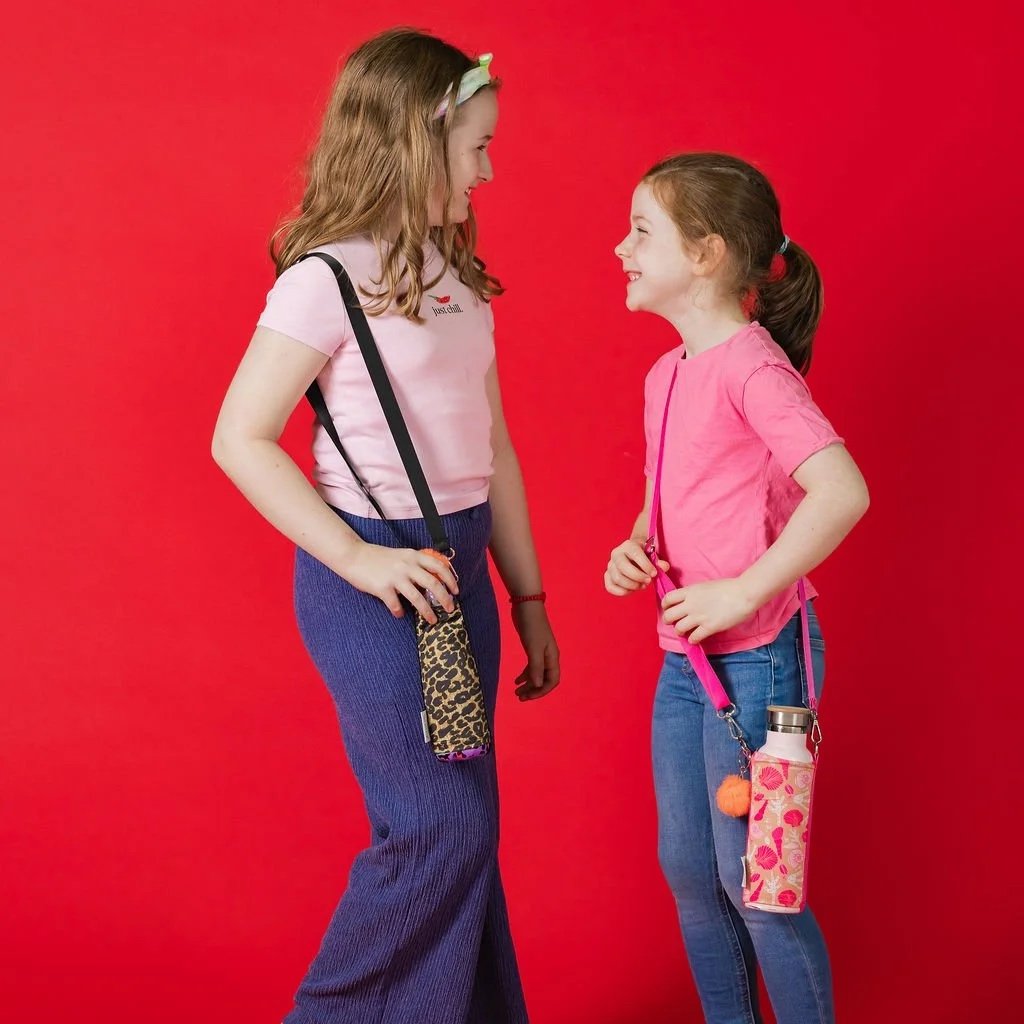 “Two girls laughing together against a red backdrop, each wearing a BottleSoc crossbody water bottle sleeve.”