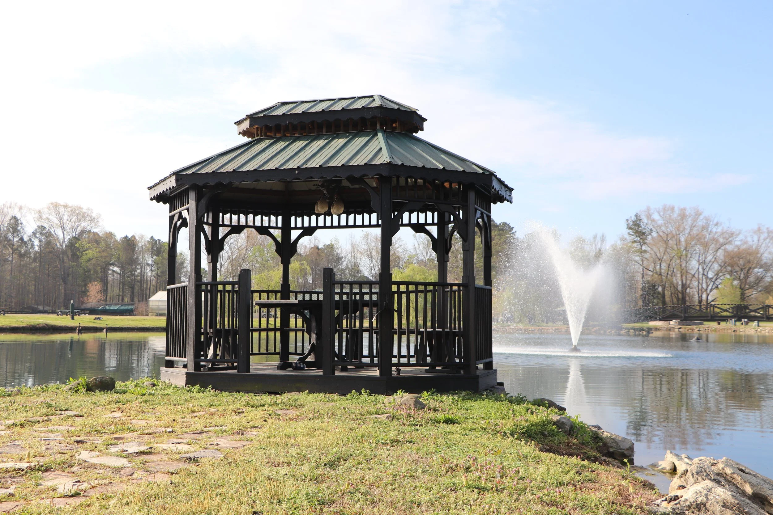 A black gazebo with a metal roof by a pond, with a fountain in the background and trees surrounding the area.