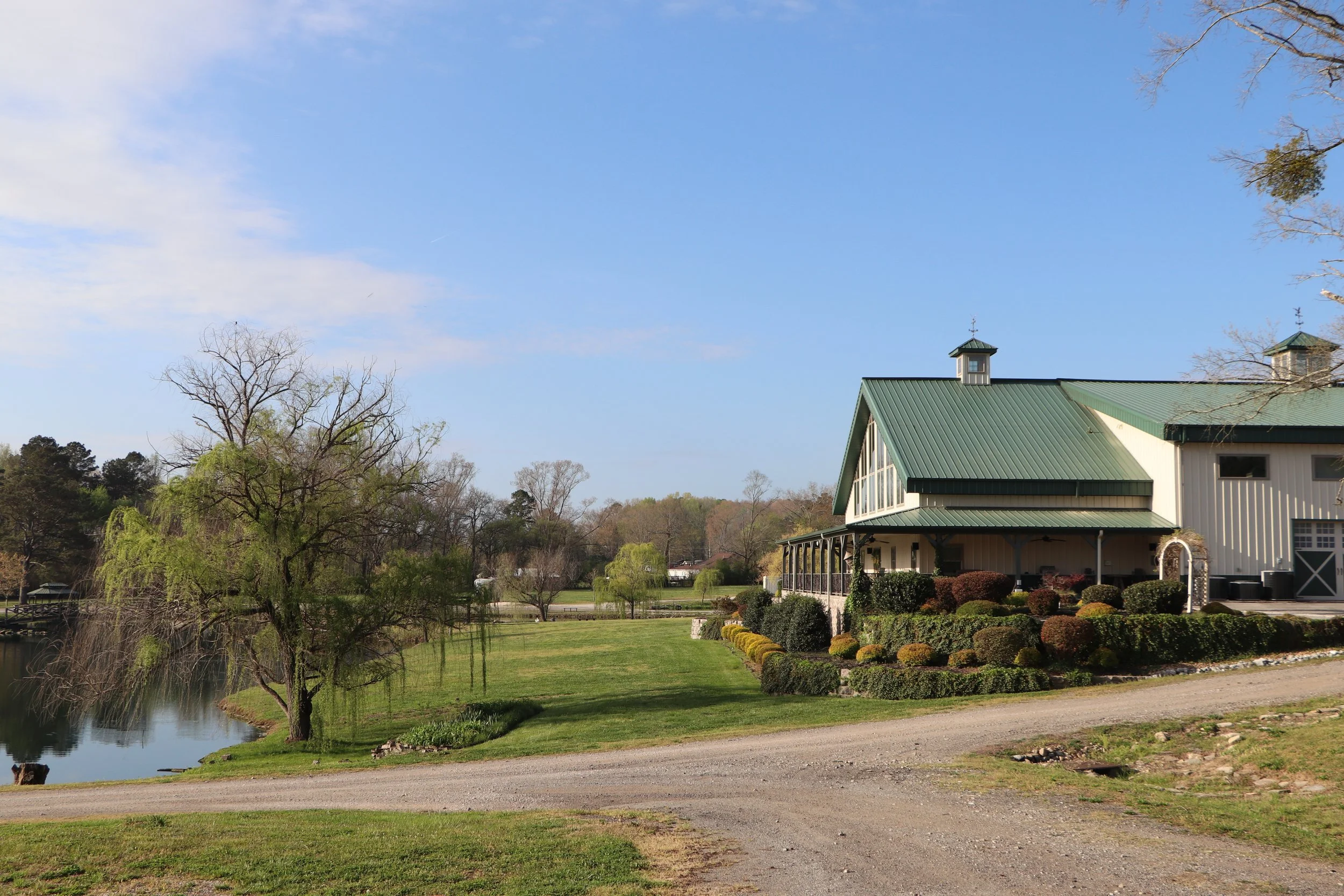 A large building with a green metal roof, surrounded by trees, shrubs, and a pond, under a clear blue sky.