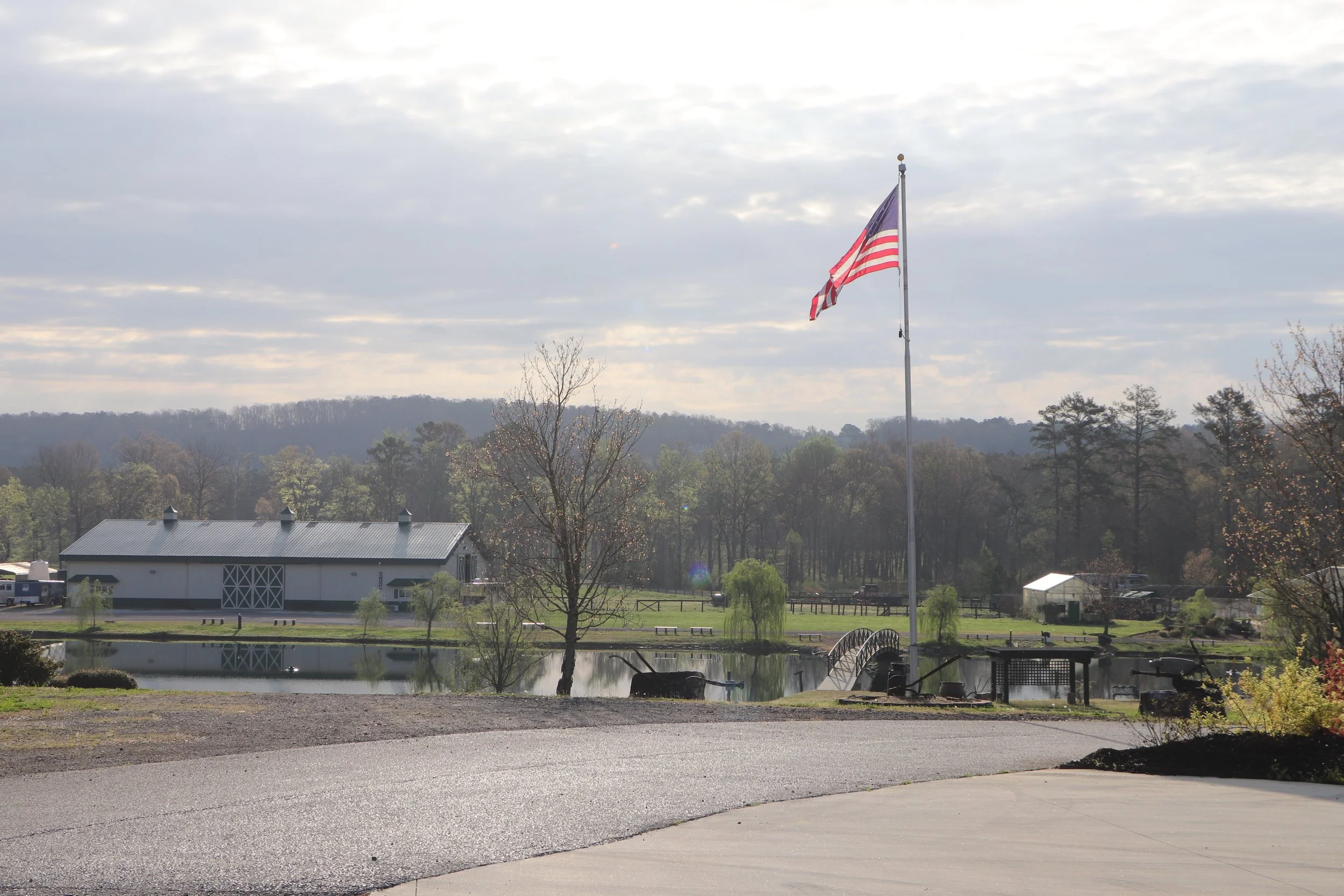 Flagpole with American flag near a pond, surrounded by trees and a white barn, cloudy sky overhead.