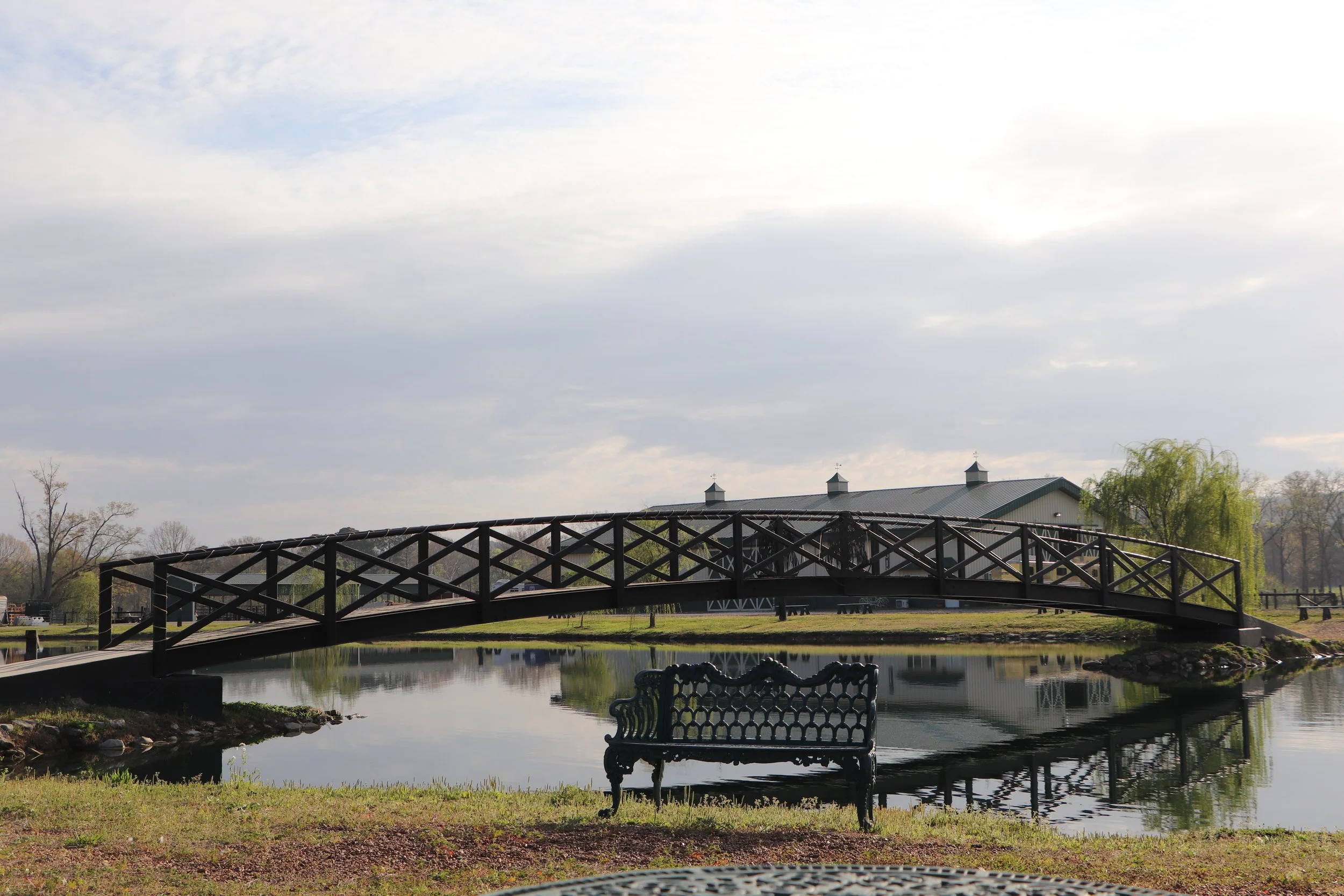 A park scene with a wooden bridge over a pond, a bench in the foreground, and a building with a metal roof and spires in the background under a cloudy sky.
