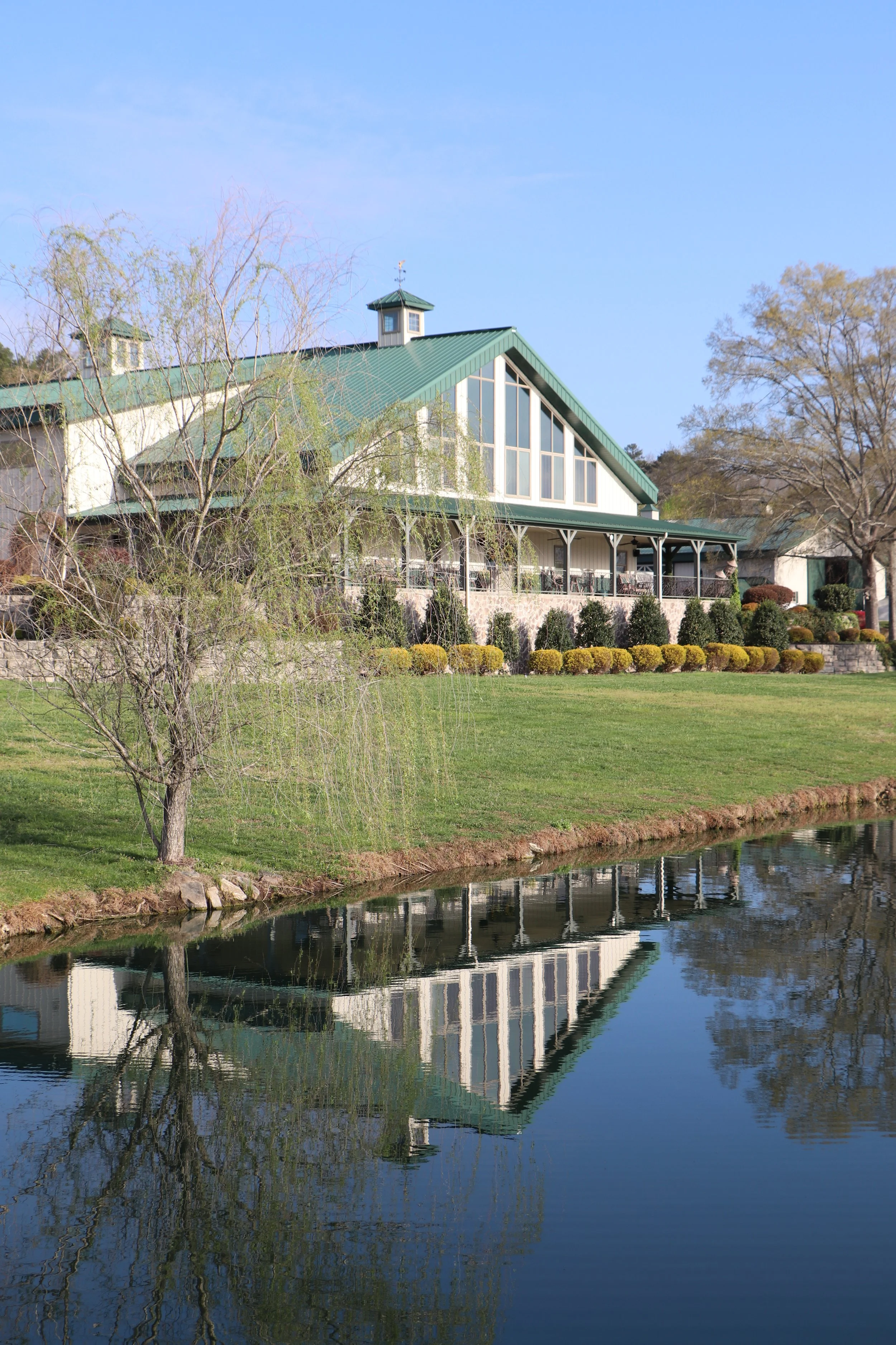 A large white house with a green metal roof and large glass windows, situated by a small pond, with trees and bushes in the yard.