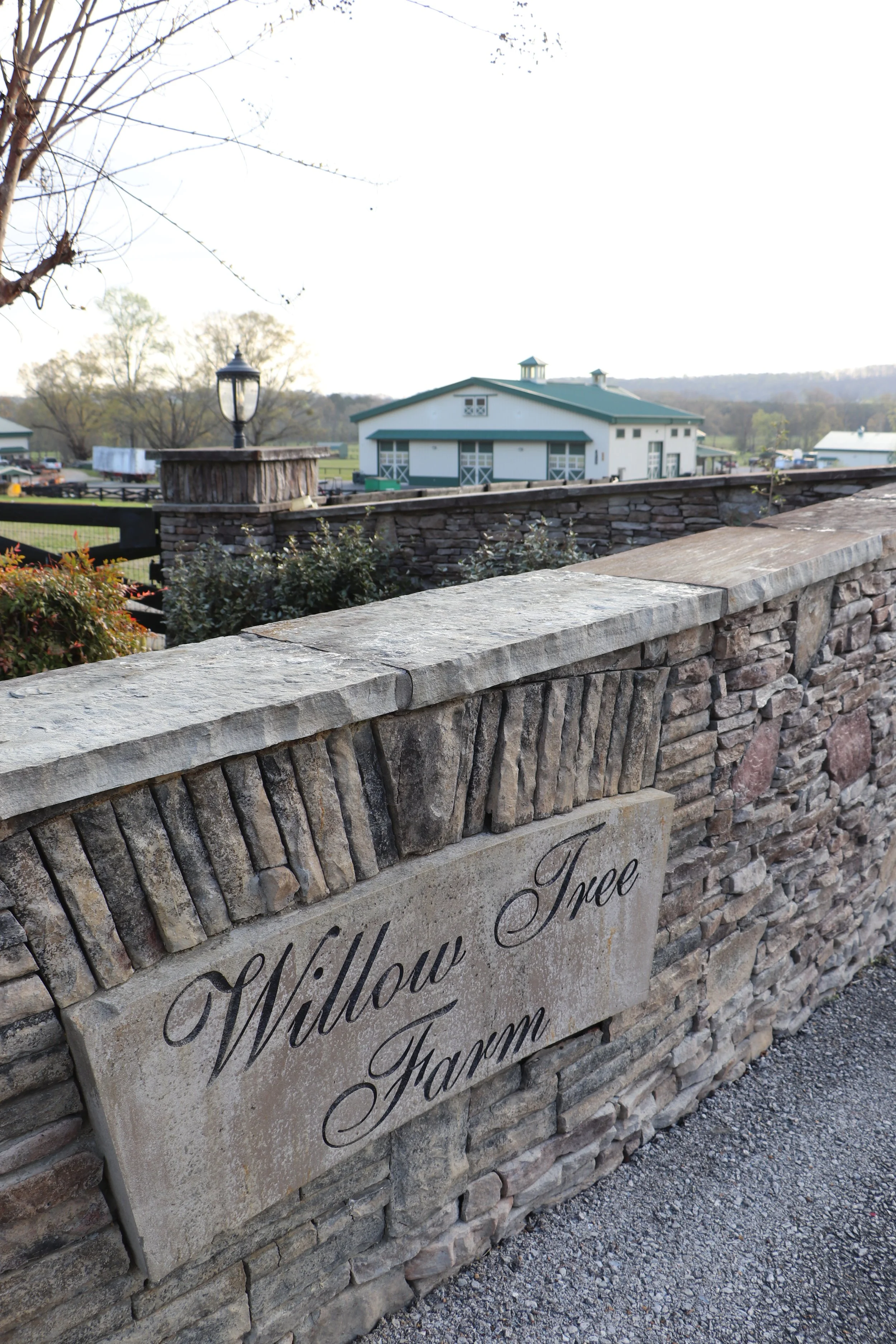 A stone sign with black cursive writing that reads "Willow Valley" and "Willow Farm" in the foreground, a stone wall, a black lamp post, and a white farm building with green trim and a metal roof in the background. There are leafless trees and a farm