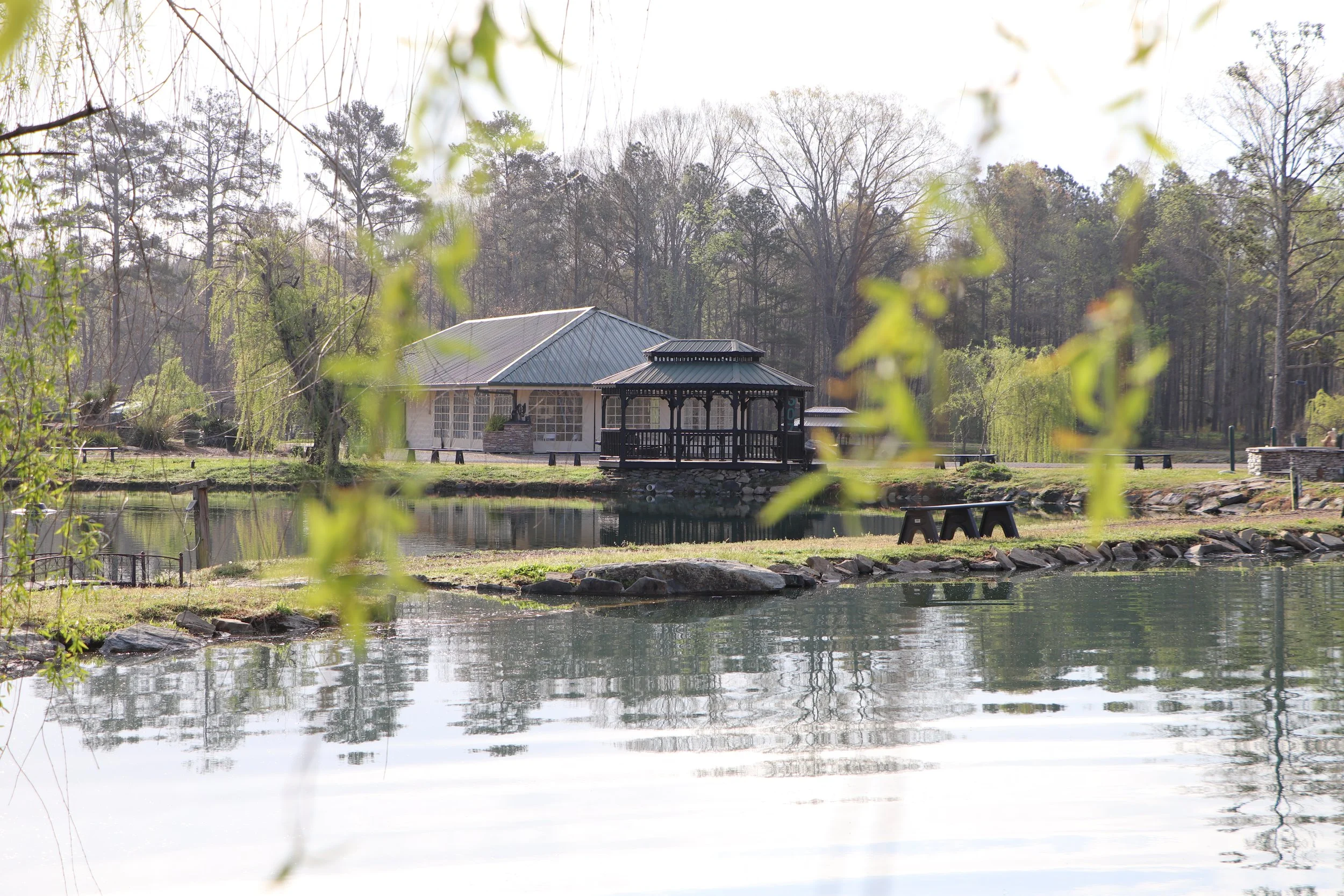 A serene lakeside scene with a small wooden gazebo on the water, surrounded by trees and a building in the background, with reflections on the calm water surface.