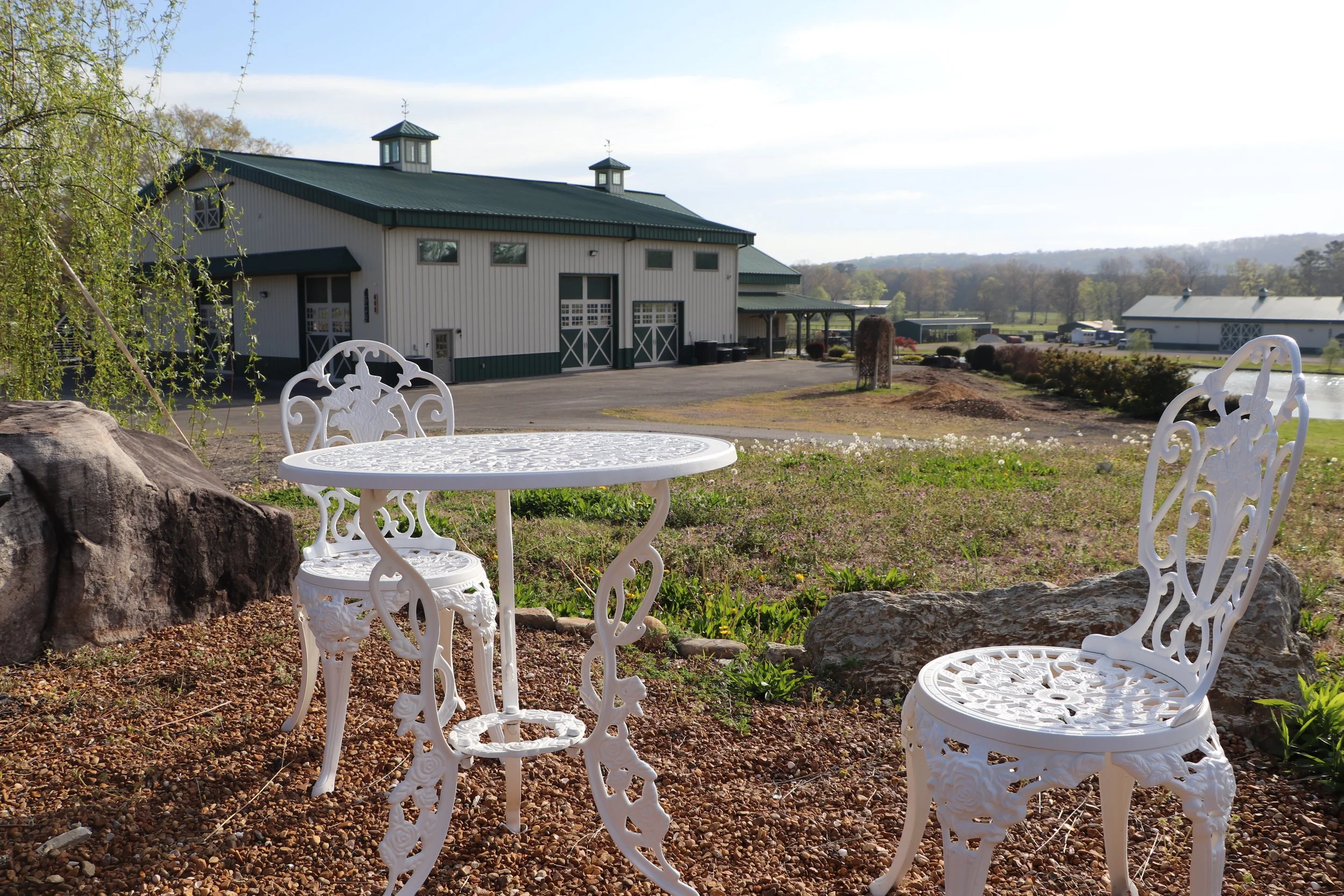 A rural landscape with white ornate metal garden furniture, a large barn with a green roof, and rolling hills in the distance under a partly cloudy sky.