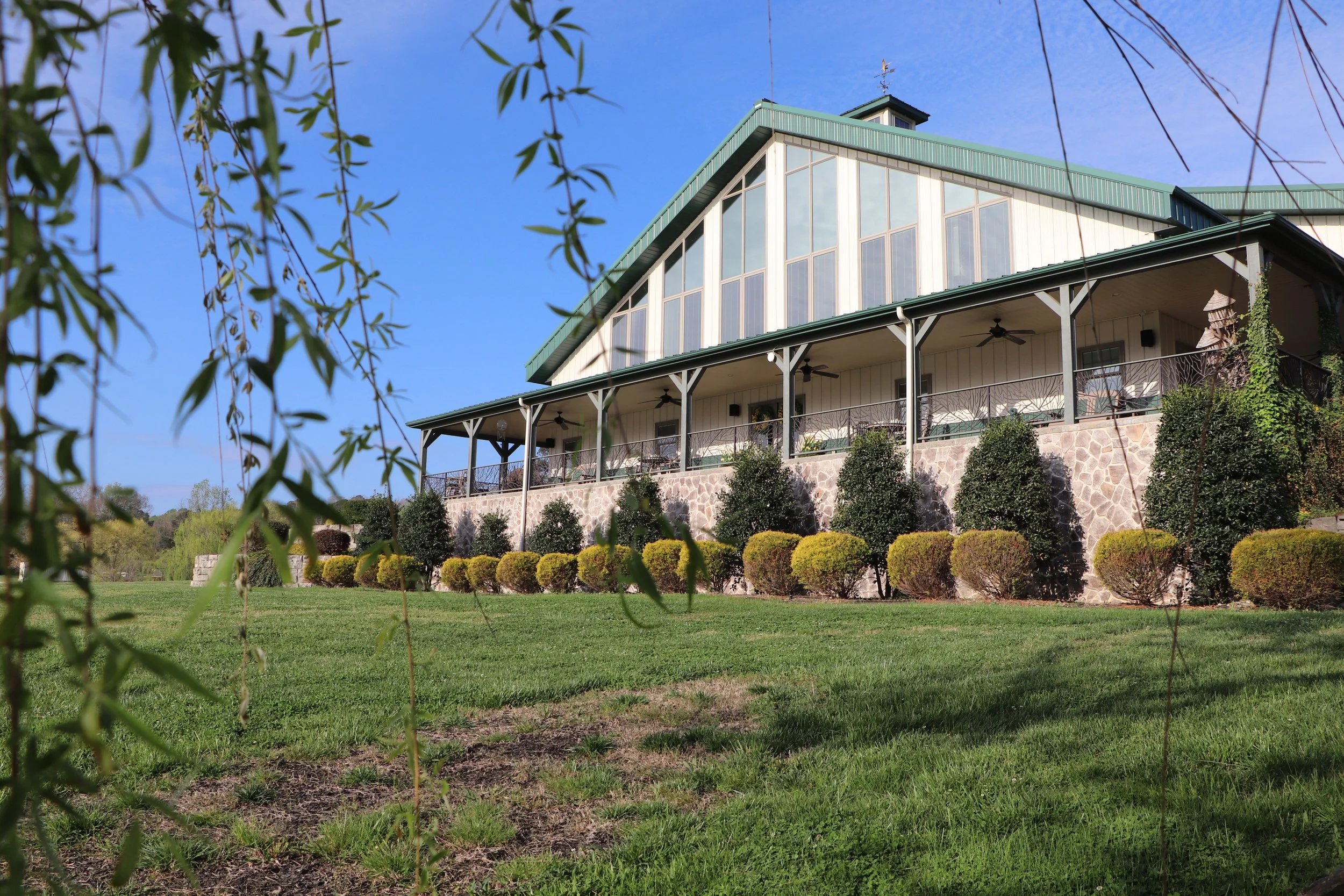 Large house with green roof and extensive windows, built on a stone foundation, surrounded by bushes and trees in a lush yard.