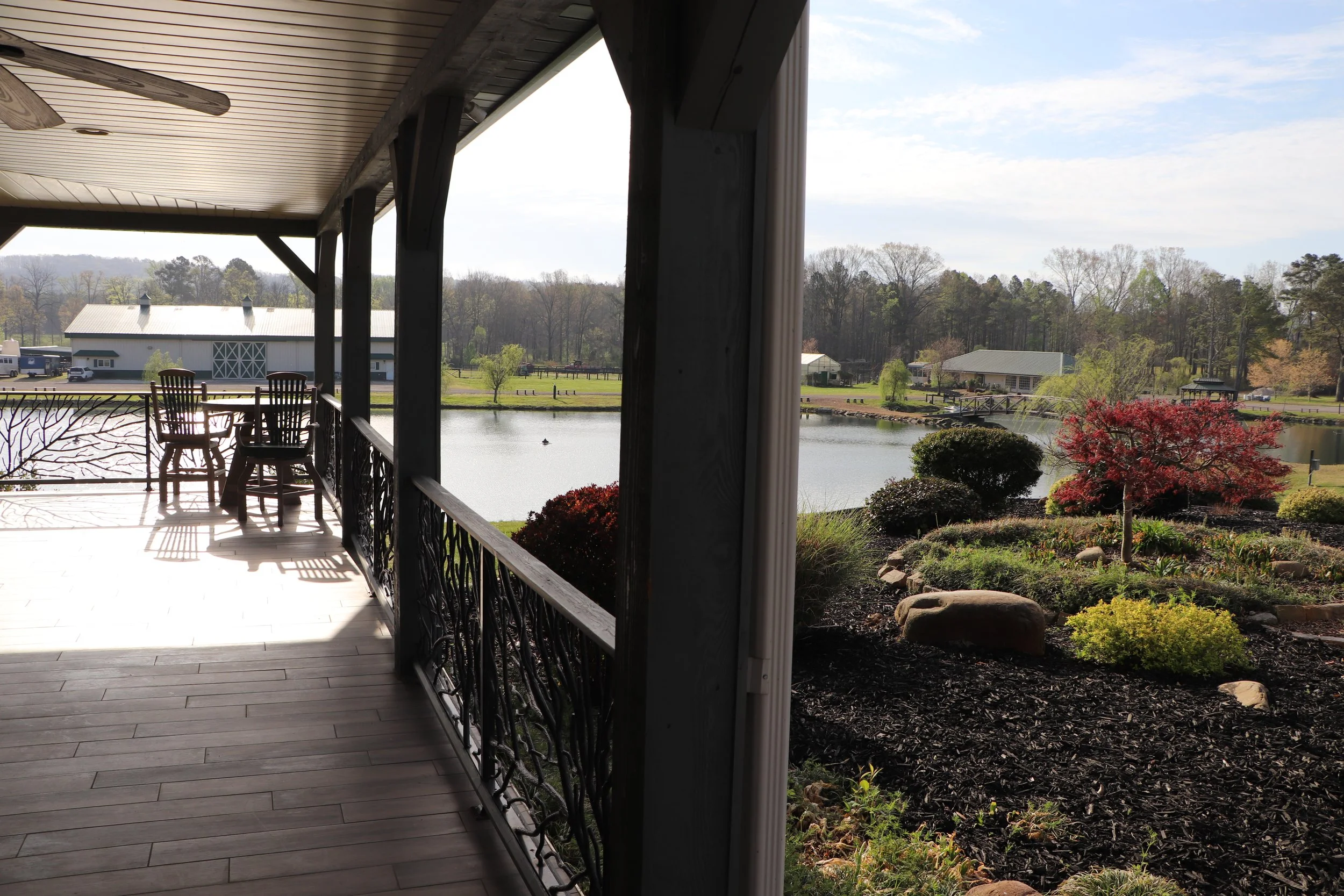 Back porch overlooking a lake with trees and buildings in the distance, wooden chairs on the porch, and a garden with a small red tree in the foreground.