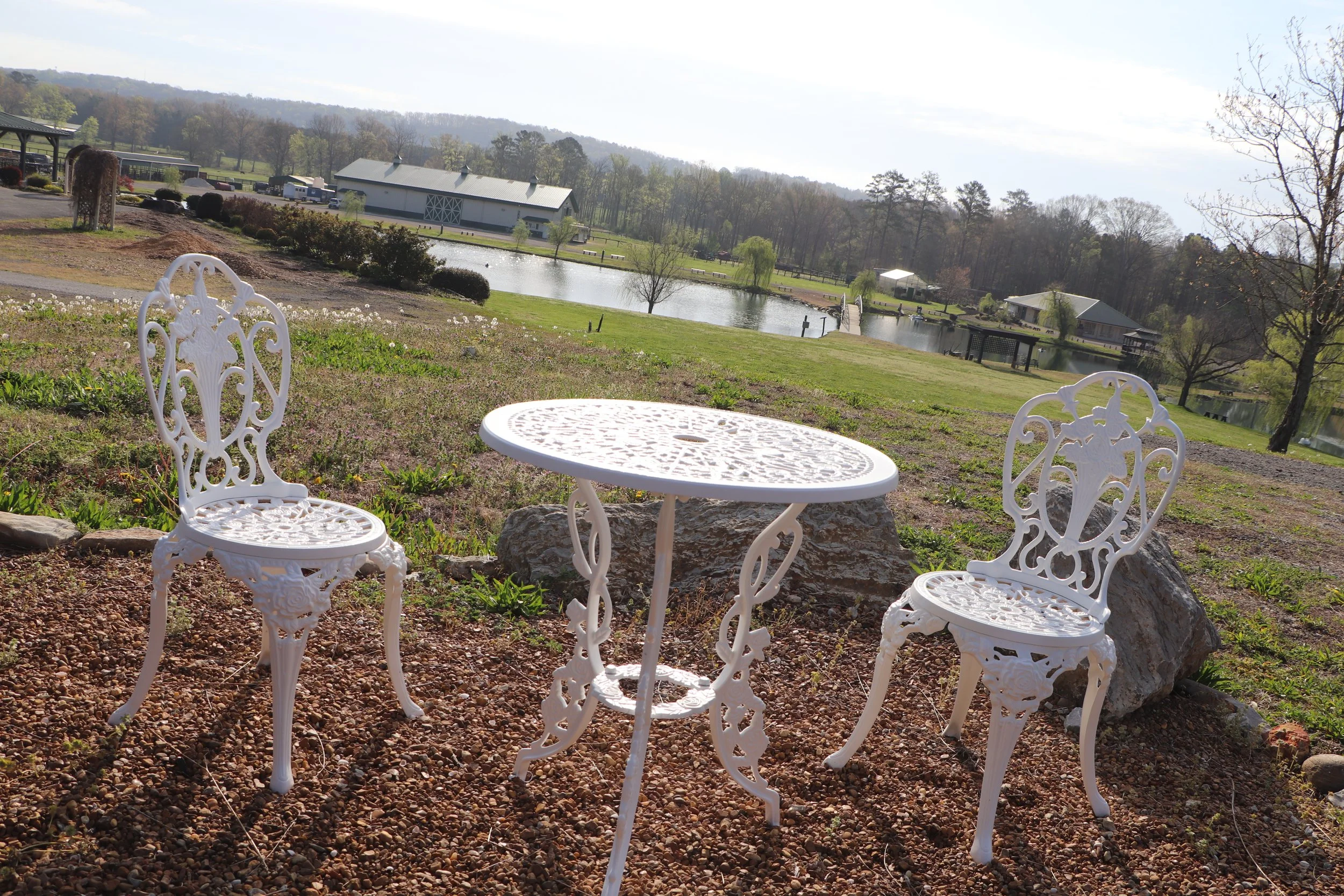 White wrought iron table and two matching chairs on gravel ground with a scenic view of a pond, trees, and buildings in the background.