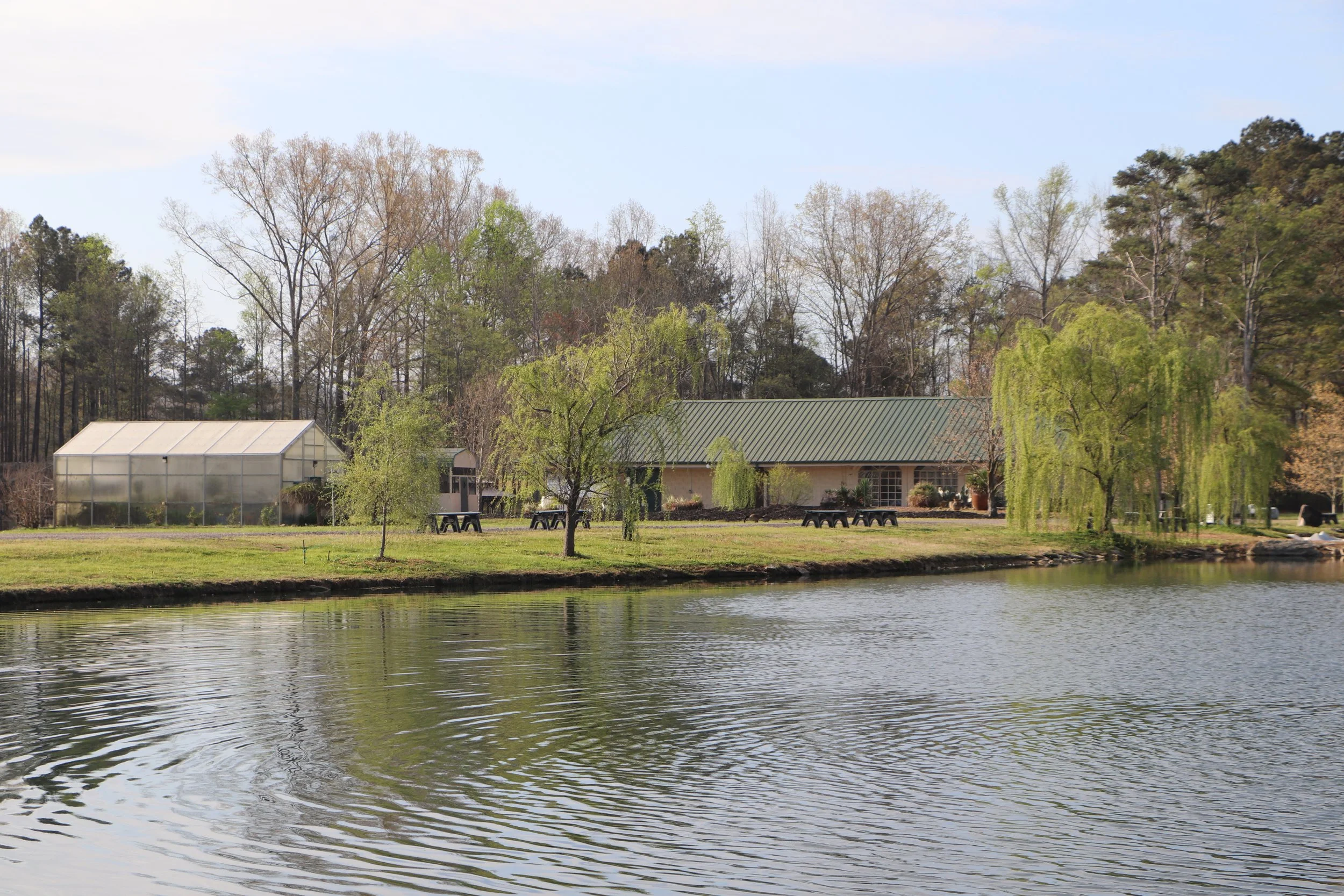 A peaceful lakeside scene with a house, greenhouses, and trees in the background, reflected on the calm water.