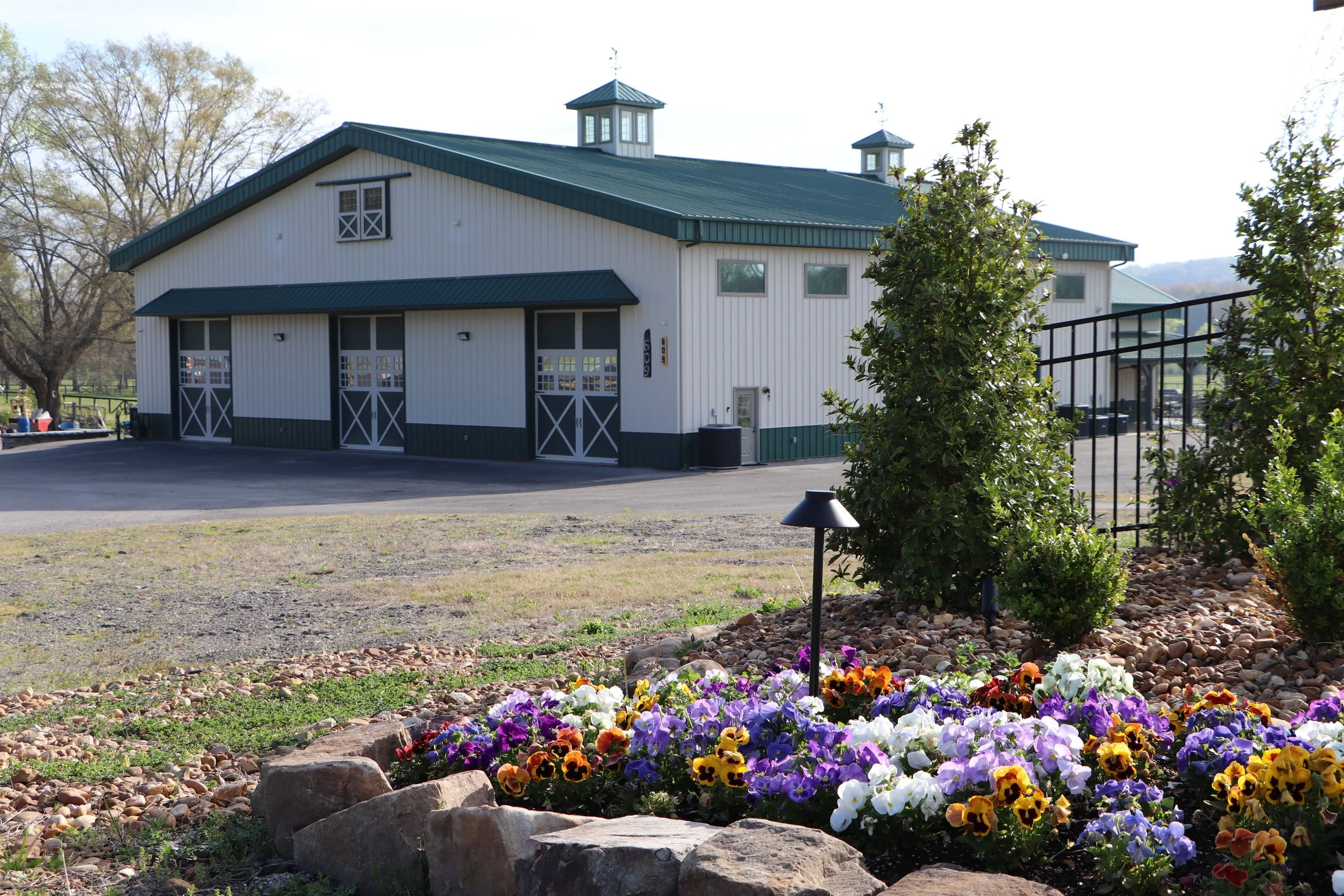 A white barn with green roof and trim, surrounded by a garden with colorful pansies and shrubs, and trees in the background.