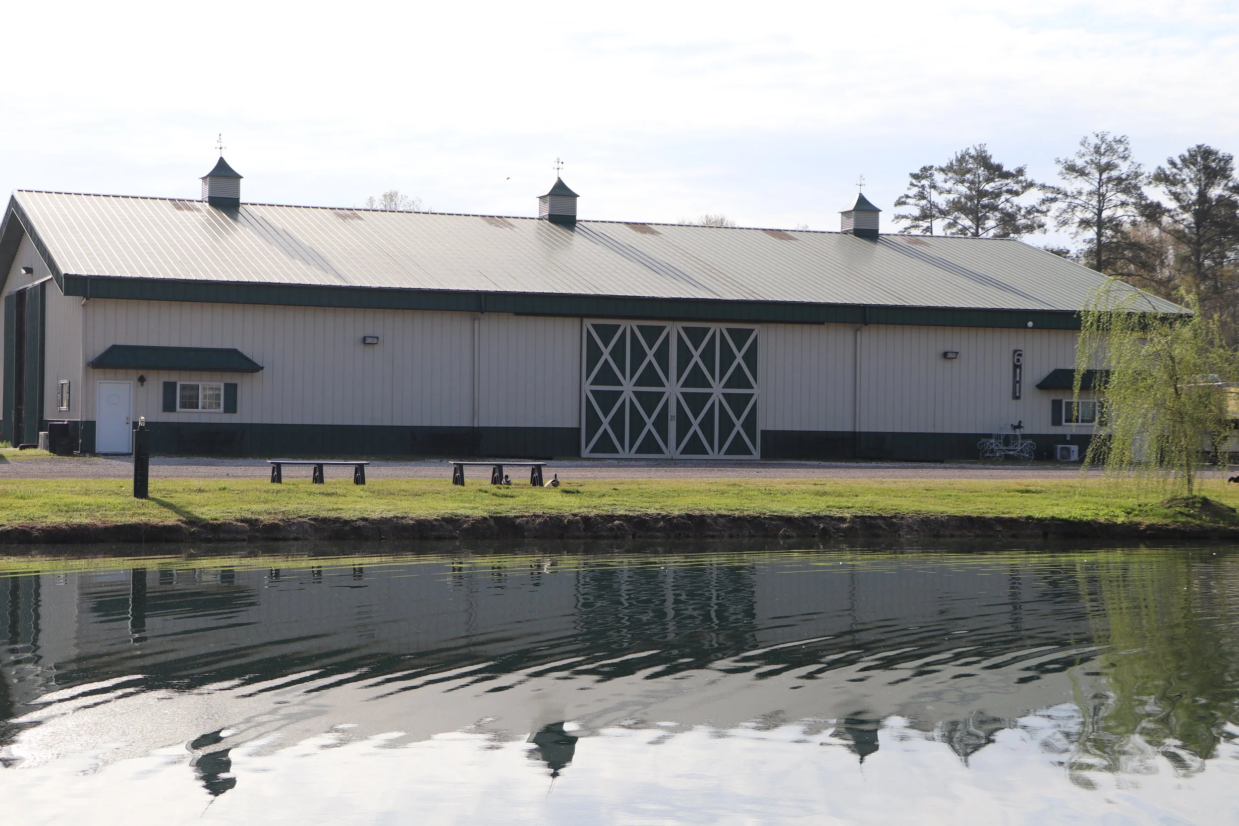 A large white barn with a metal roof and three small cupolas, situated near a body of water with trees in the background.