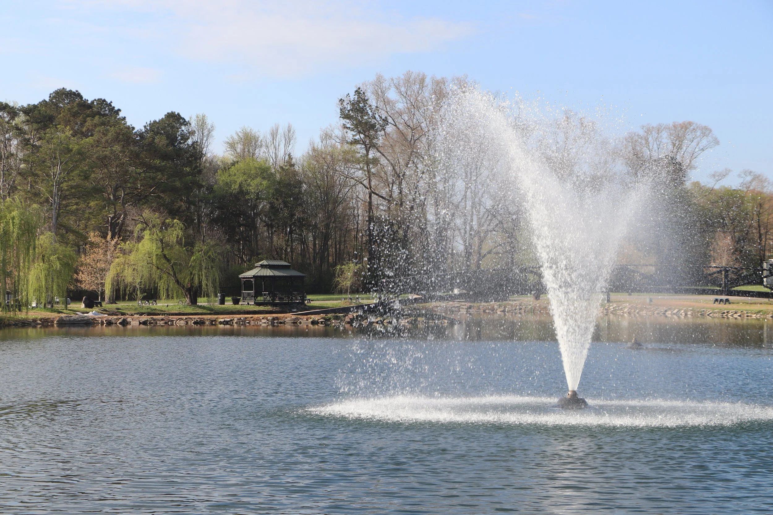 A park with a lake, trees, a small gazebo, and a fountain spraying water into the air on a sunny day.