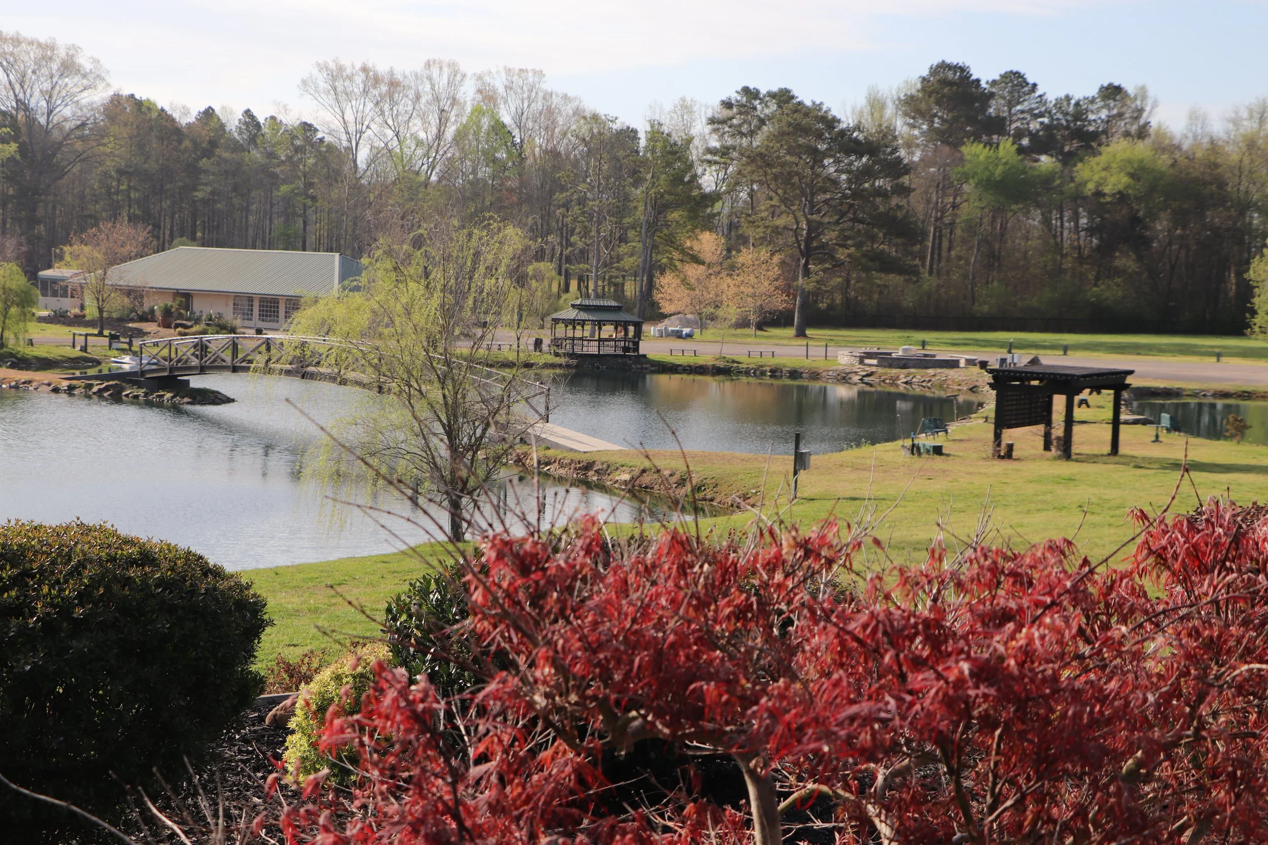 A scenic outdoor view with a pond in the foreground, a red shrub in the lower part, and a gazebo on the pond's edge. There is a building with a green roof in the background amidst trees and a partly cloudy sky.