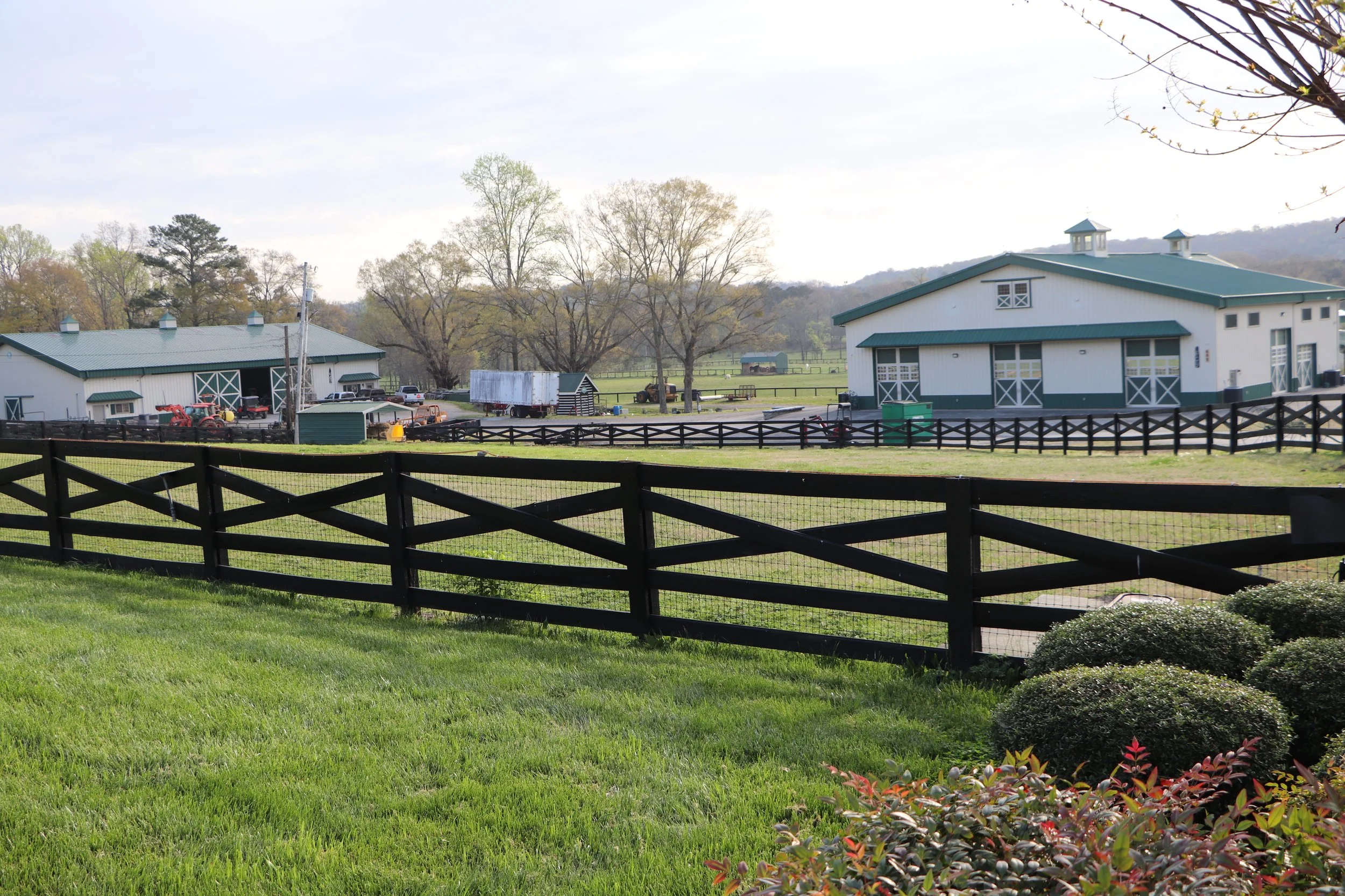 A view of a farm with green fields, two large white barns with green roofs, a black wooden fence, and trees in the background, under a partly cloudy sky.