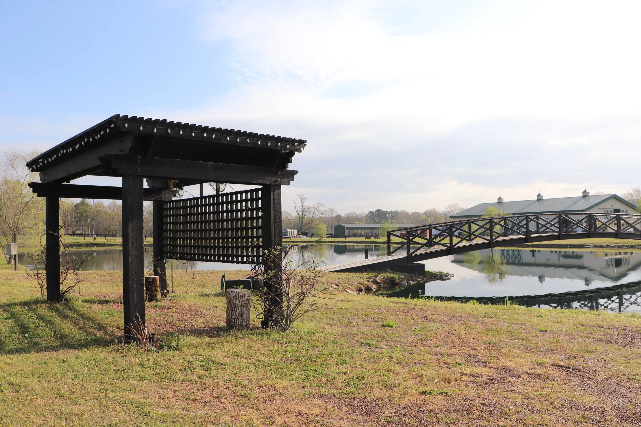 A wooden bridge over a calm pond with a building and trees in the background under a partly cloudy sky.
