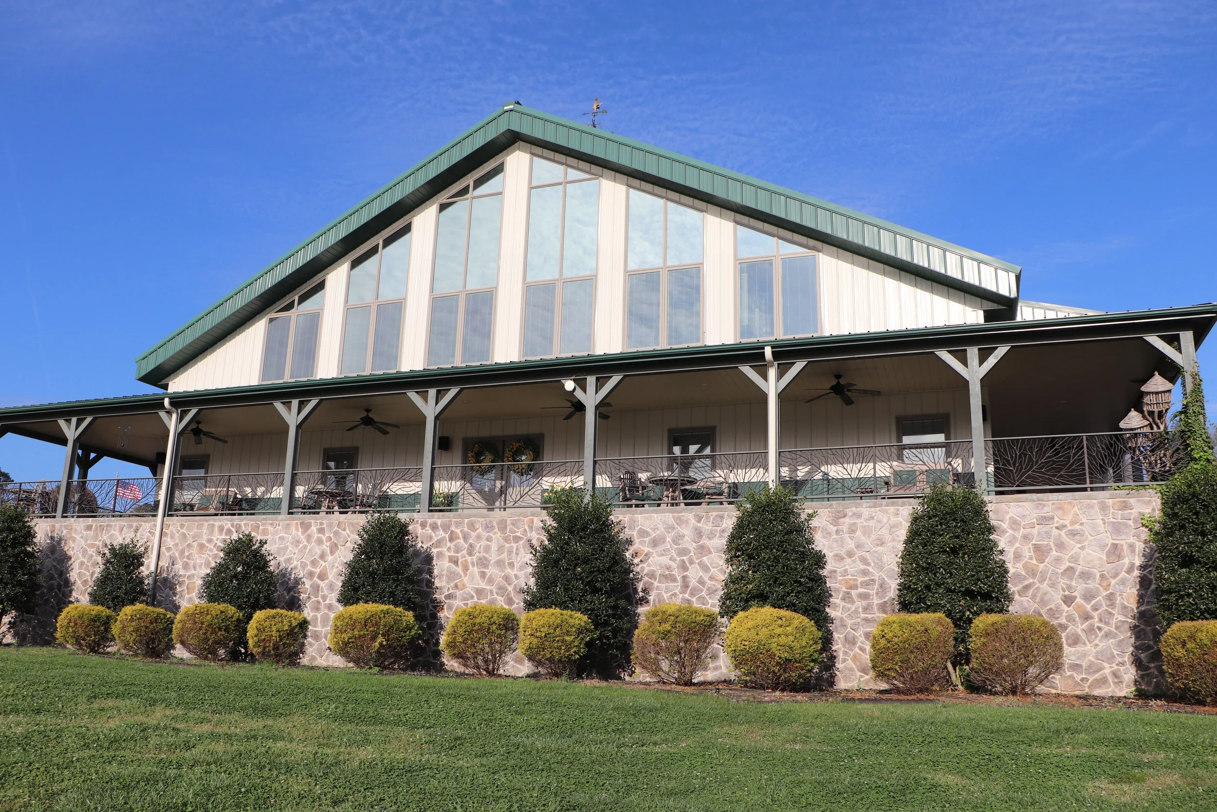 A house with large windows, a green roof, a stone foundation, and a balcony with outdoor furniture and decorations, surrounded by a well-maintained lawn and shrubs, under a clear blue sky.