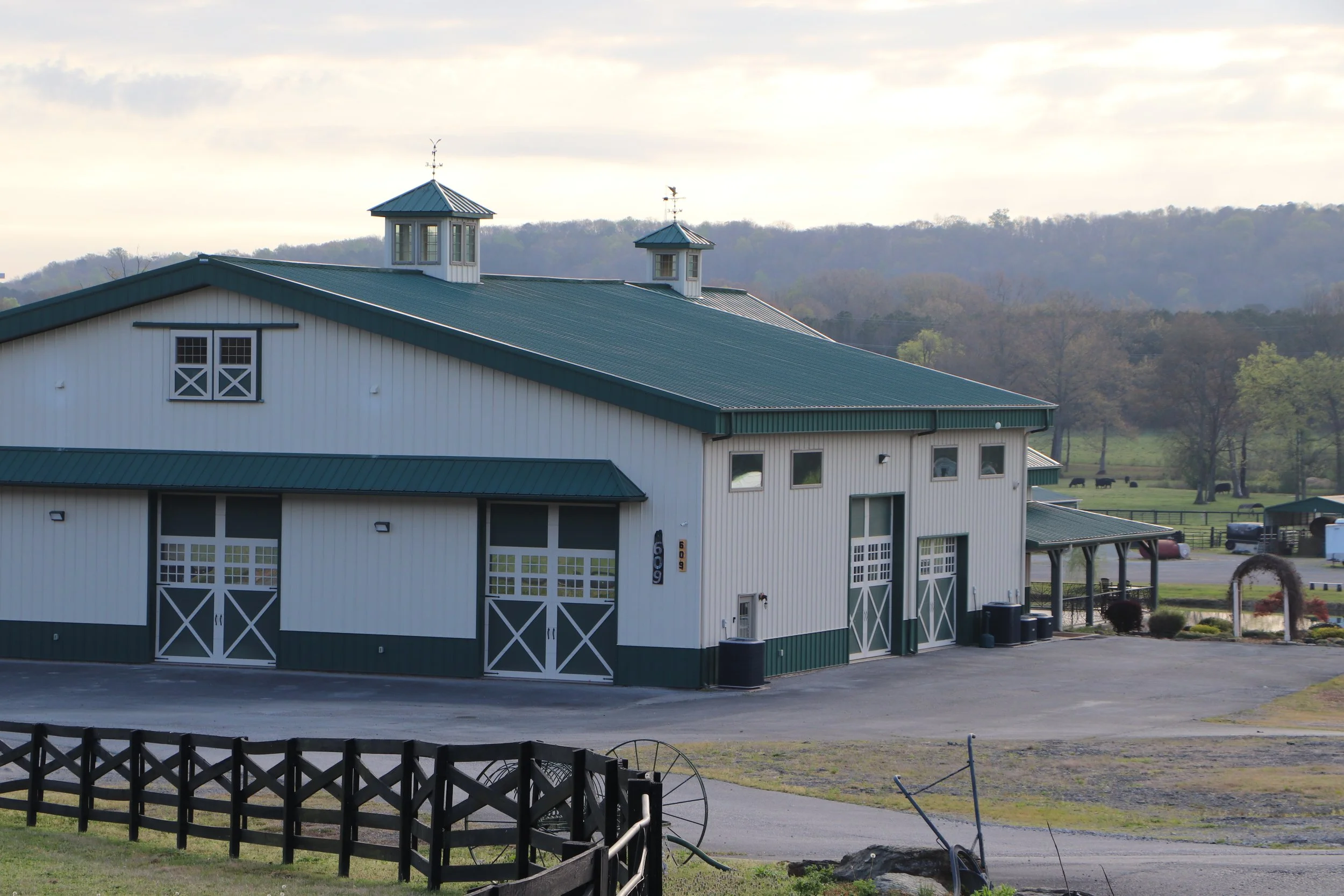 A large white barn with a green metal roof, located in a rural area with hills and trees in the background. The barn has two small cupolas on the roof with weather vanes, and multiple garage-style doors on the front with white trim. There is a black 