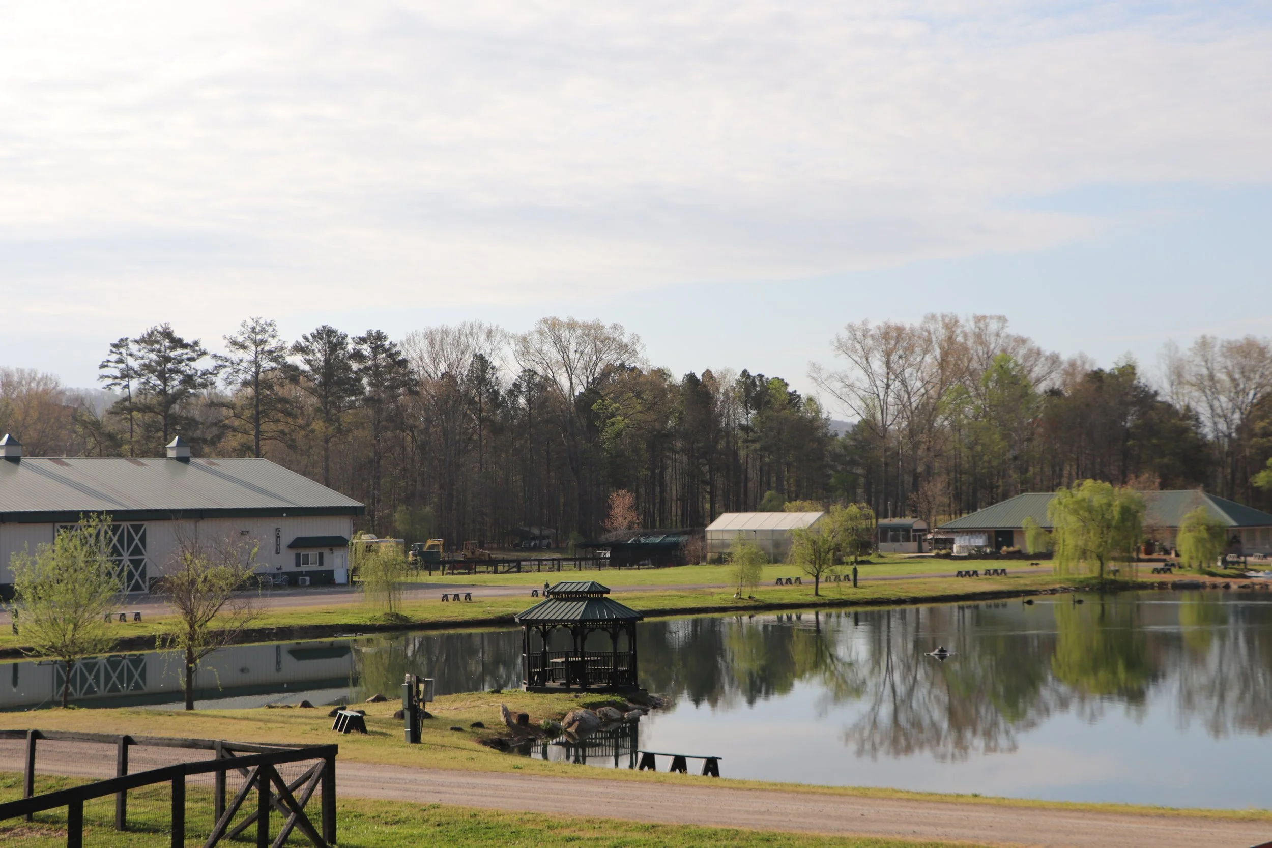 A scenic view of a pond with a small gazebo, surrounded by green trees, some buildings with green roofs, picnic tables, and a cloudy sky overhead.