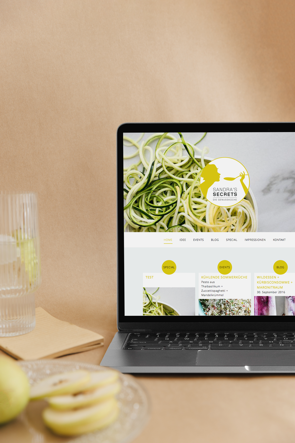 Ein Laptop zeigt eine Webseite namens "Sandra's Secrets" mit einem Bild von Zucchini-Nudeln und einem Menü mit Kategorien wie Home, Ideen, Events, Blog, Spezial, Impressionen, Kontakt. Im Vordergrund sind eine halbe grüne Frucht, eine Glasschale mit 