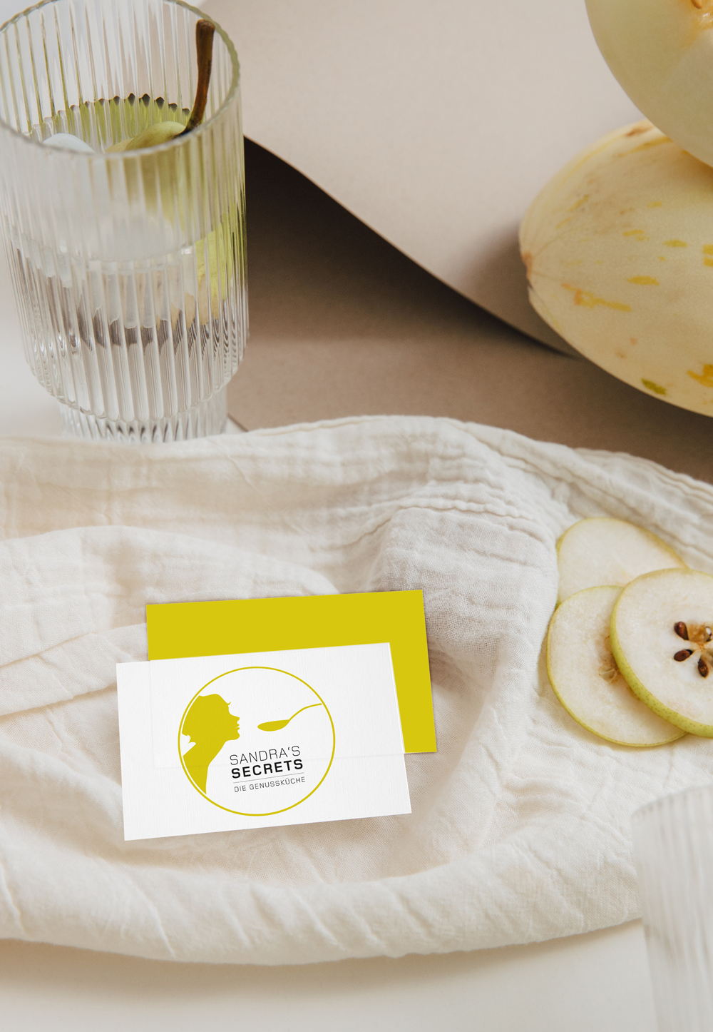 Eine weiße Stoffserviette mit einem weißen und grünen Apfel, zwei Apfelscheiben, ein Glas Wasser mit Apfel und Zimtstange, eine Kartennummer von Sandra's Secrets.