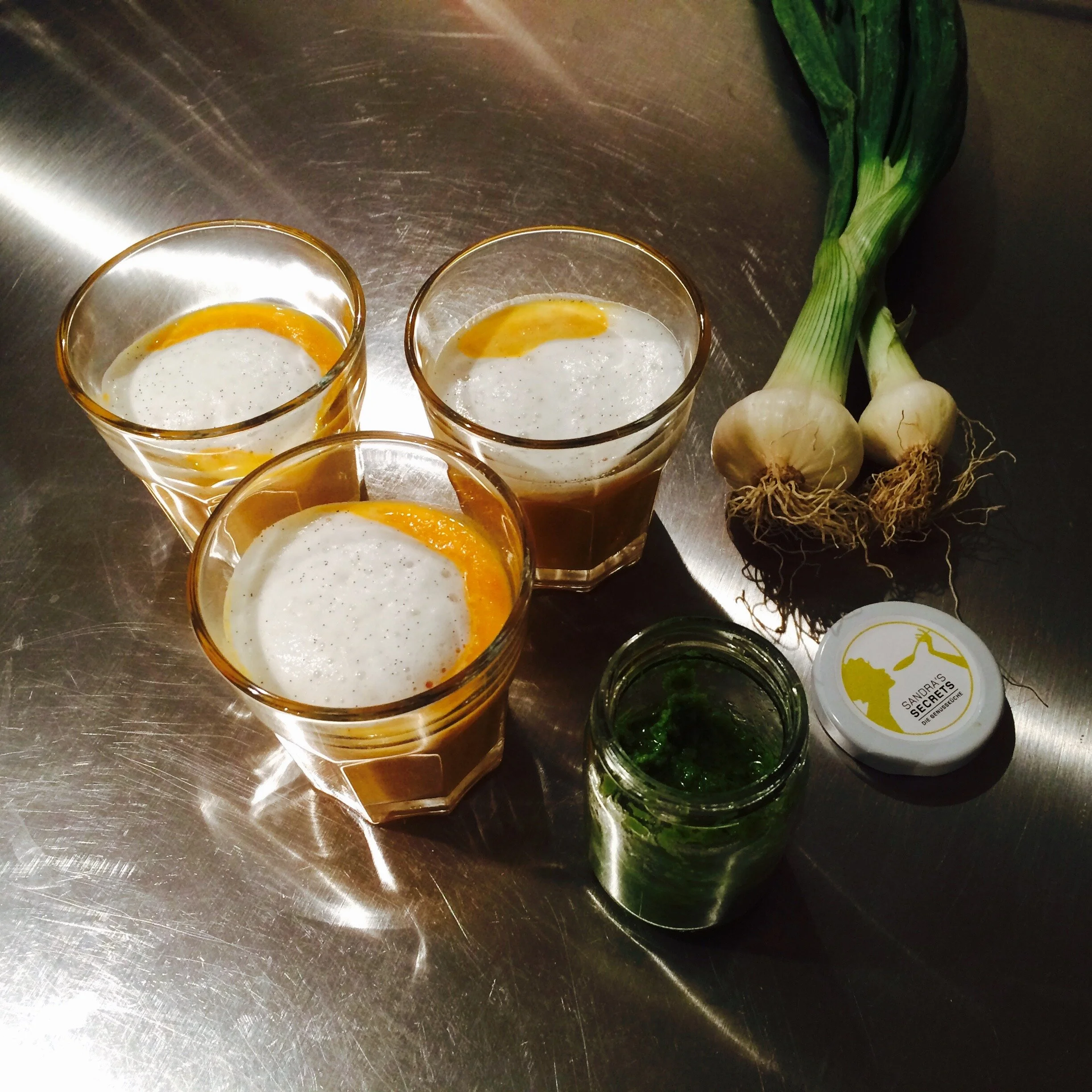 Drei Gläser mit Getränken, Knoblauch, ein kleiner Behälter mit grünen Kräutern, und eine Flasche sitzen auf einer metallischen Oberfläche.