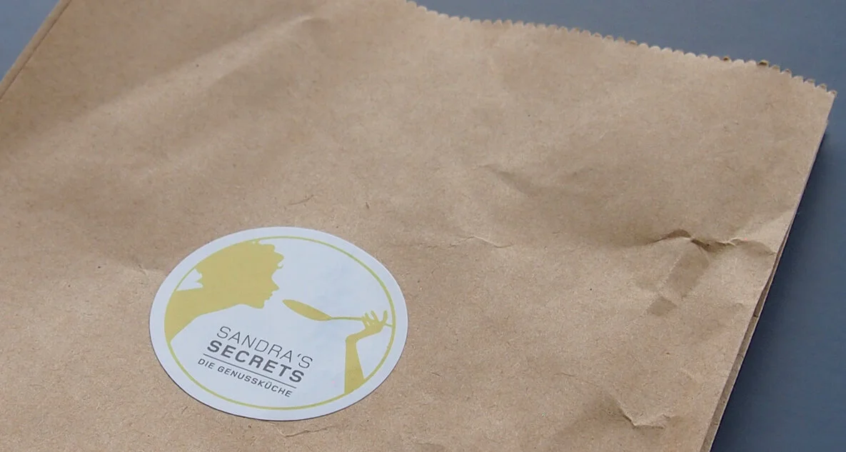 Ein brauner Papierbeutel mit einem runden Aufkleber, auf dem eine stilisierte Frau im Profil zu sehen ist, die mit einem Löffel eine Speise genießt, und die Aufschrift "Sandra's Secrets - Die Genussküche".