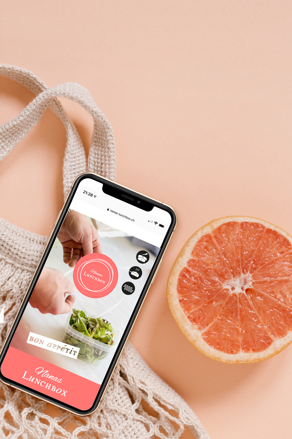 Ein Smartphone auf einem rosa Hintergrund, das eine Website mit einem Lunch-Box-Design zeigt, daneben eine halbe Grapefruit, eine weiße Tote-Tasche und ein Knoten aus Strickstoff.