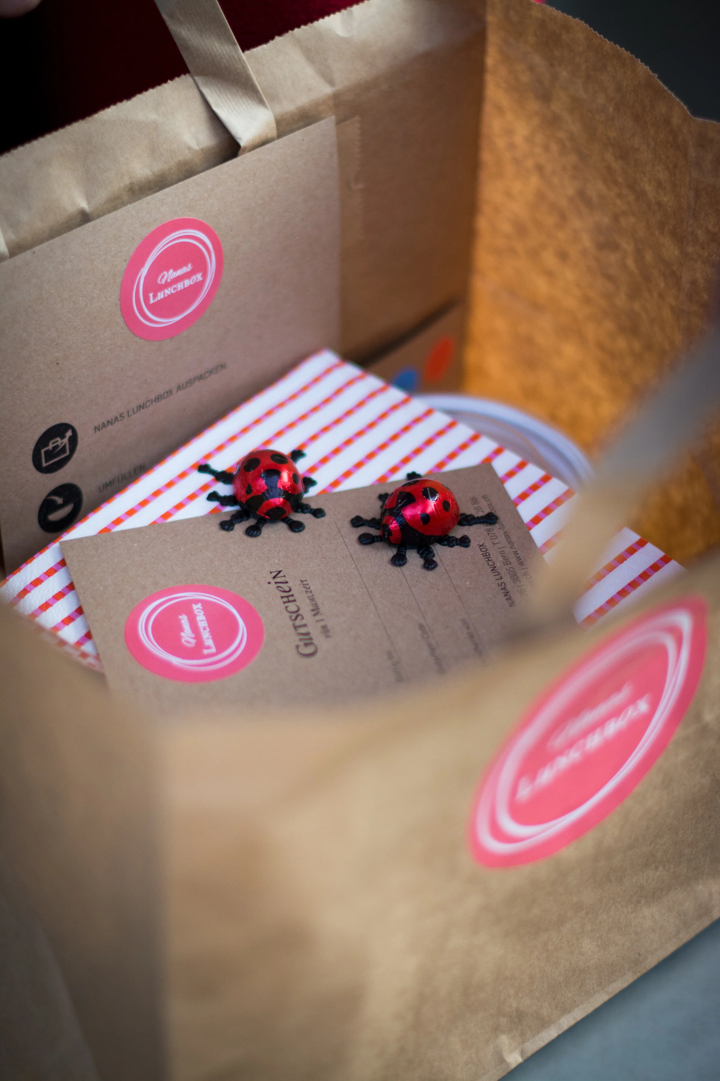 Papiertüte mit buntem Geschenkpapier, Gutschein, kleine Marienkäfer-Figuren und Lunchbox im Inneren.