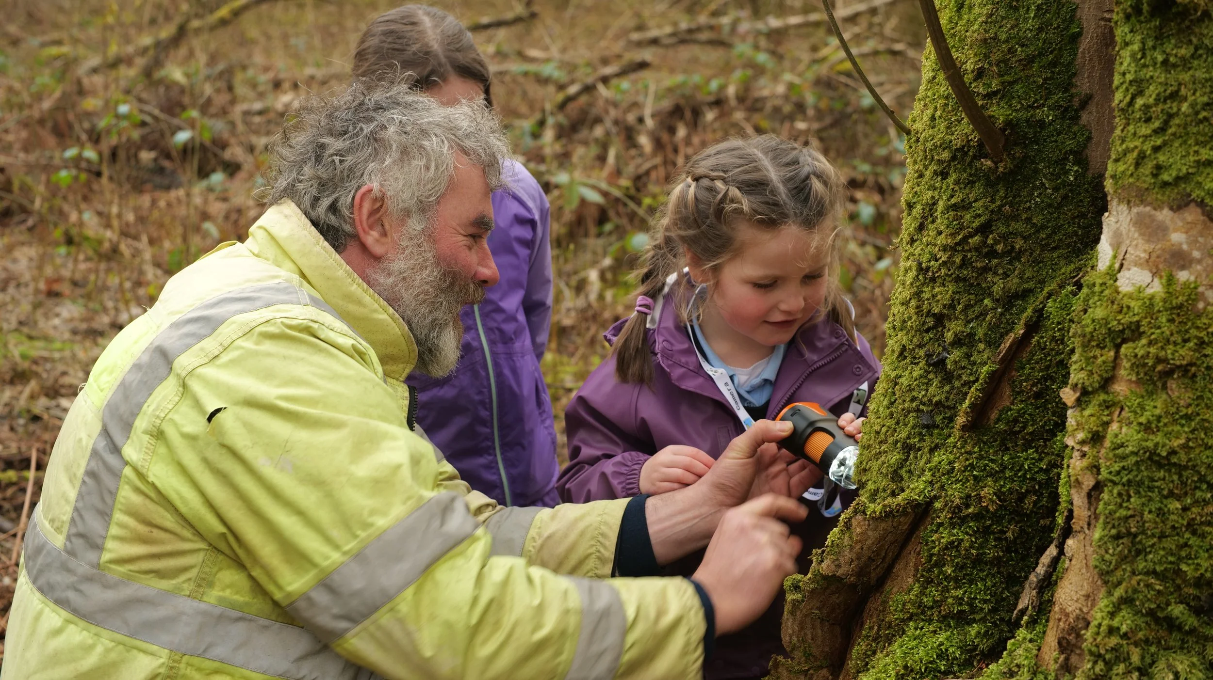 A man in a high-vis jacket shows a young girl in a purple jacket how to look at lichens through an electronic device. They're outdoors in a wet woodland