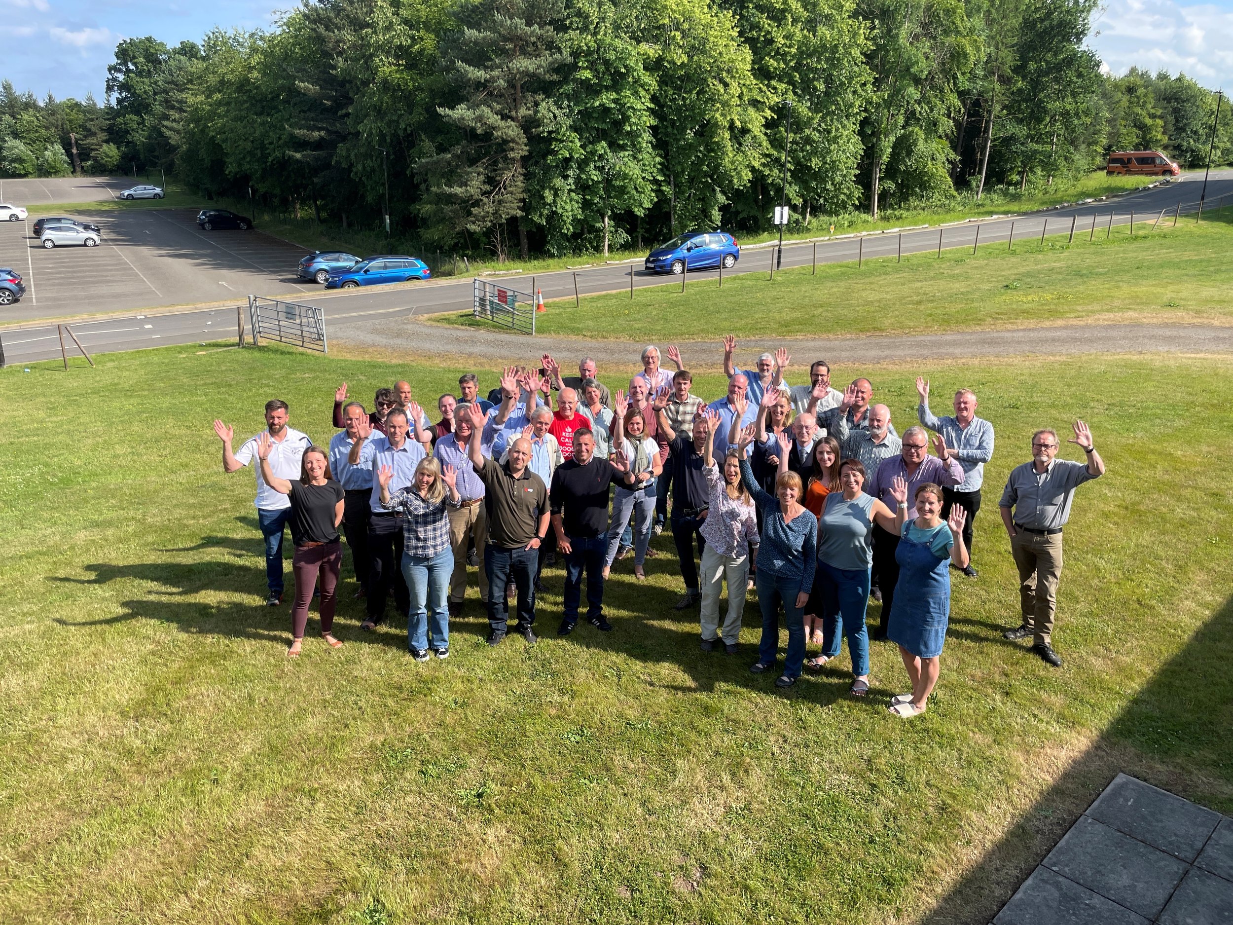 A drone shot of a group of people who are waving at the camera. It's summer and the sun is shining. They're standing on green grass with a road and trees behind them.