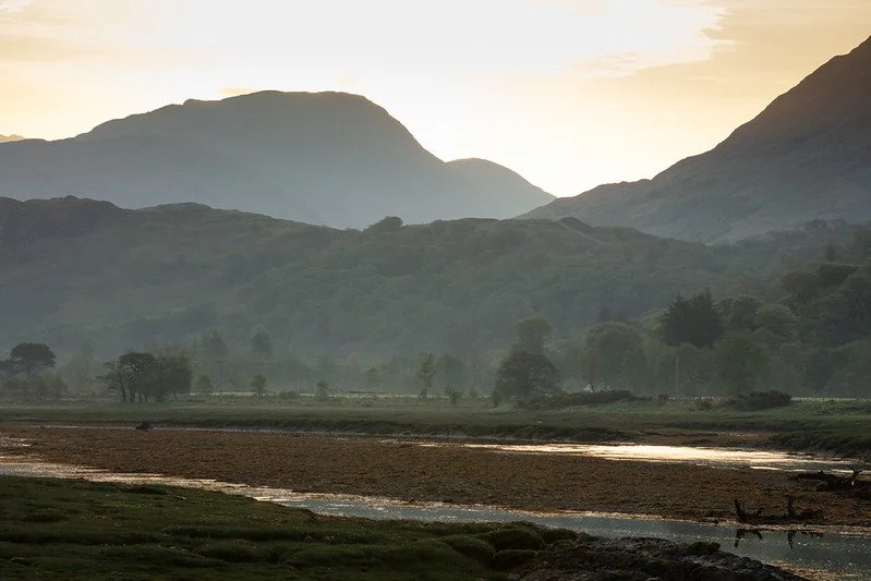 Beautiful scene of a flat river valley with the water in the foreground and trees in the distance and then mountains and the dawn sky beyond