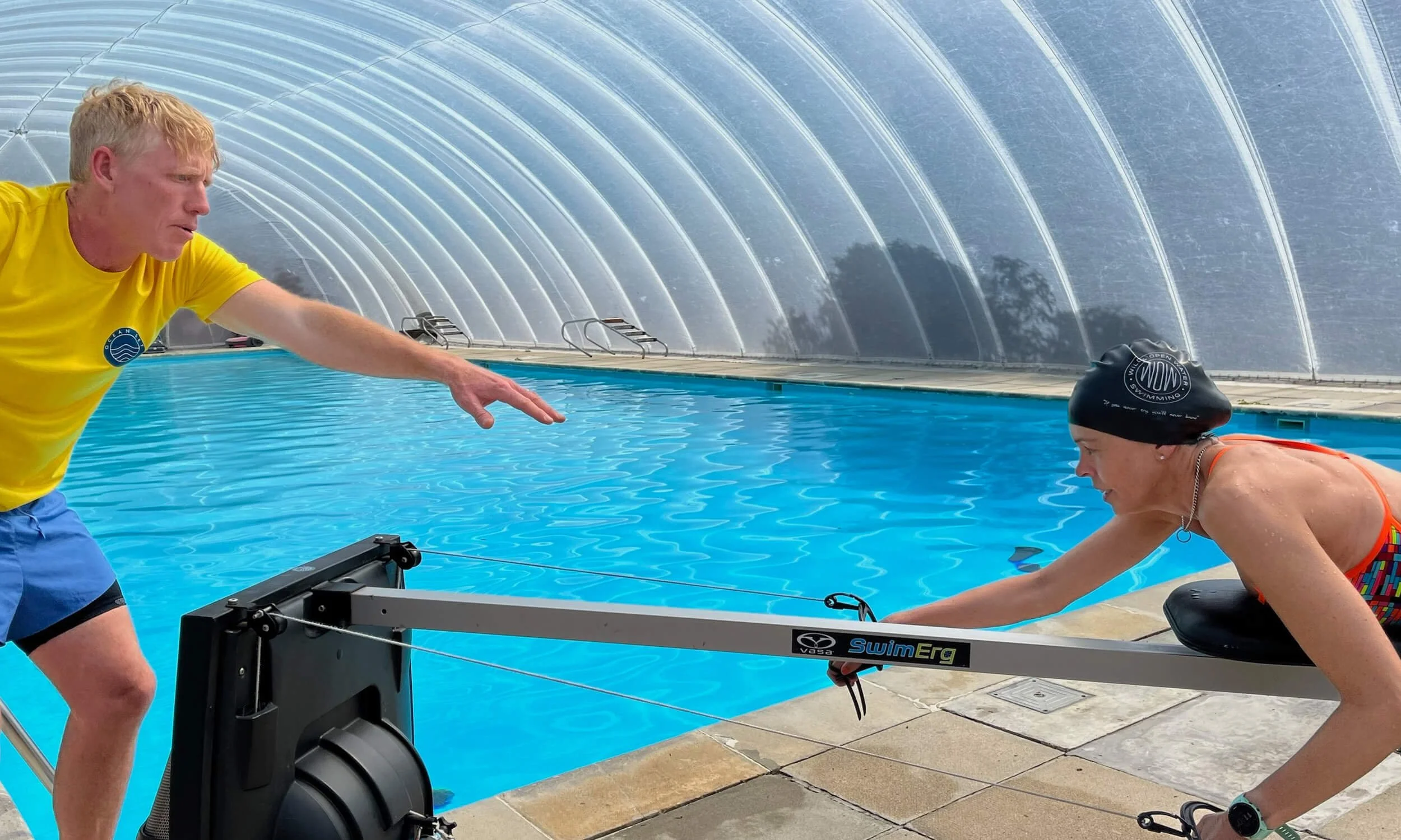 A man and a woman are exercising together by a swimming pool indoors. The man in a yellow shirt is assisting the woman who is on an exercise machine, wearing a black swim cap and colorful swimsuit. The background shows a large curved ceiling and the 