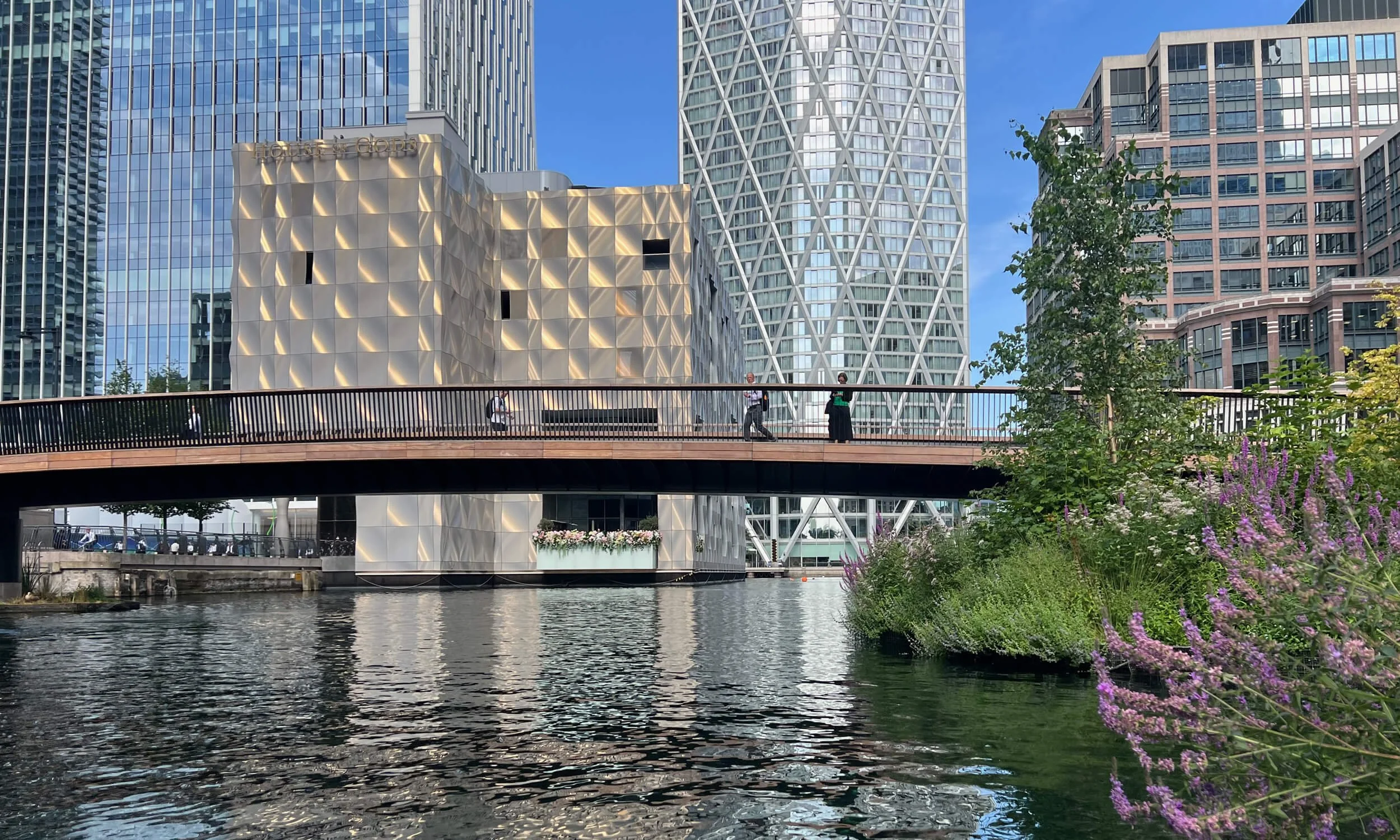 Canary Wharf cityscape featuring modern skyscrapers with reflective glass windows and a waterway in the foreground. There is a pedestrian bridge with people walking, and greenery with purple flowers on the right side of the image.