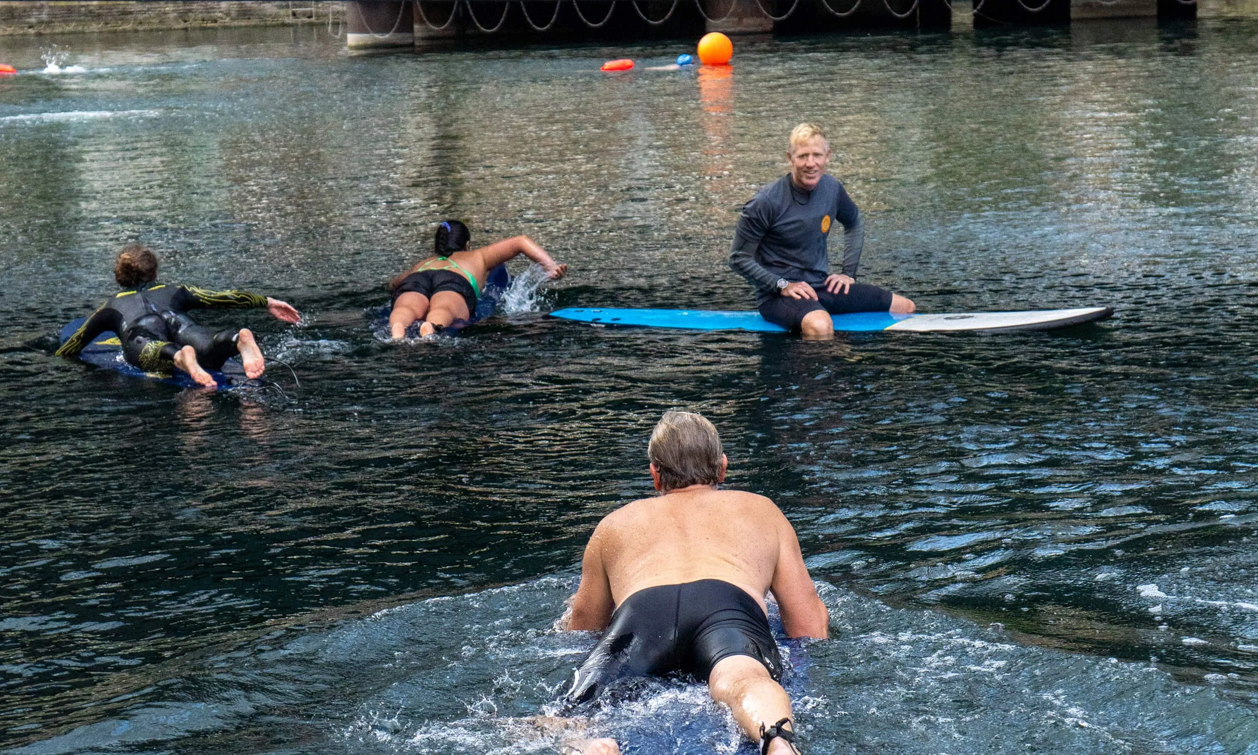 People paddling on surfboards in a body of water near a dock, with a surf coach sitting on a surfboard in the water.