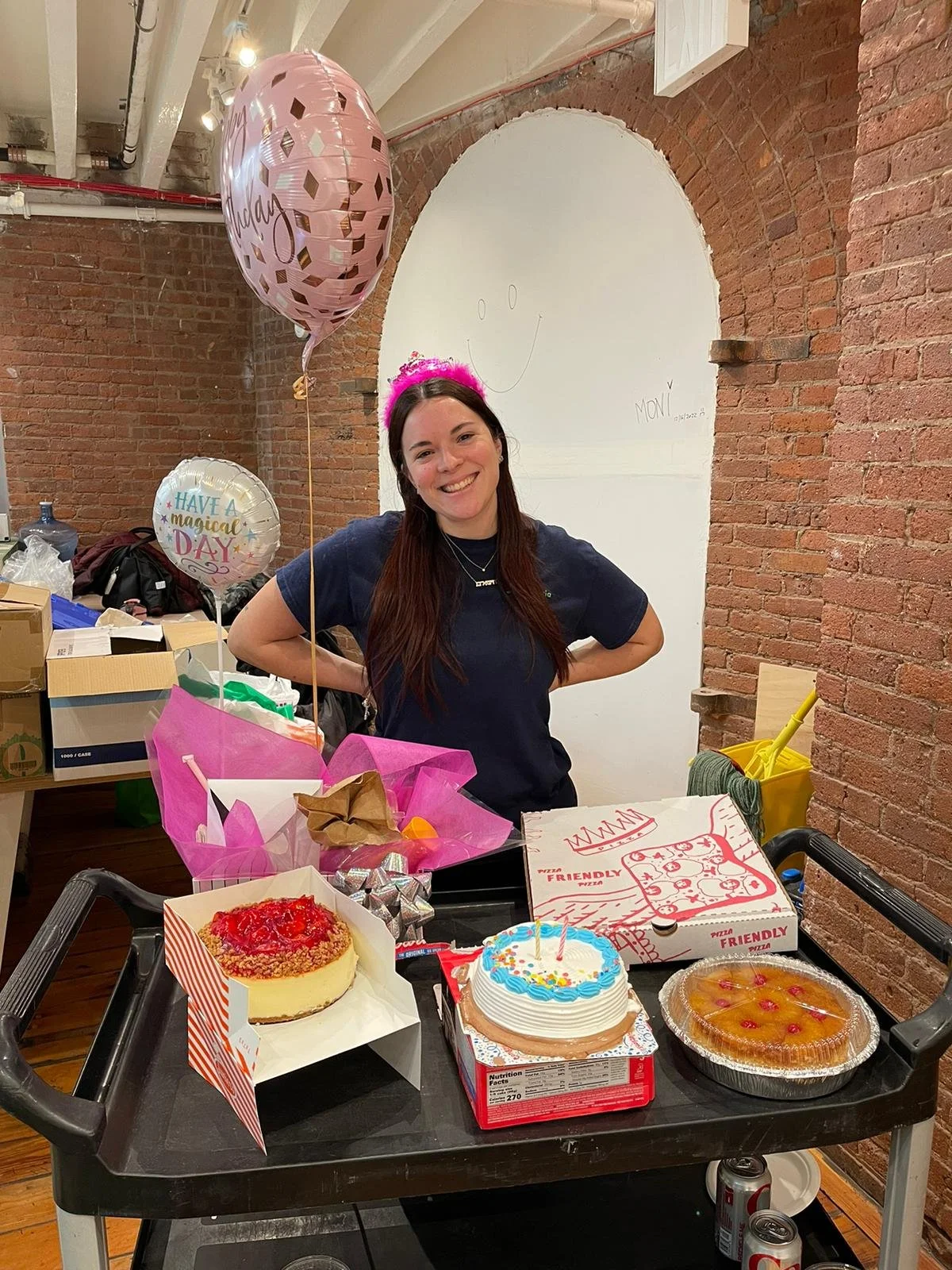 Woman with a pink birthday crown smiling at a birthday celebration with cake, pizza, and balloons.