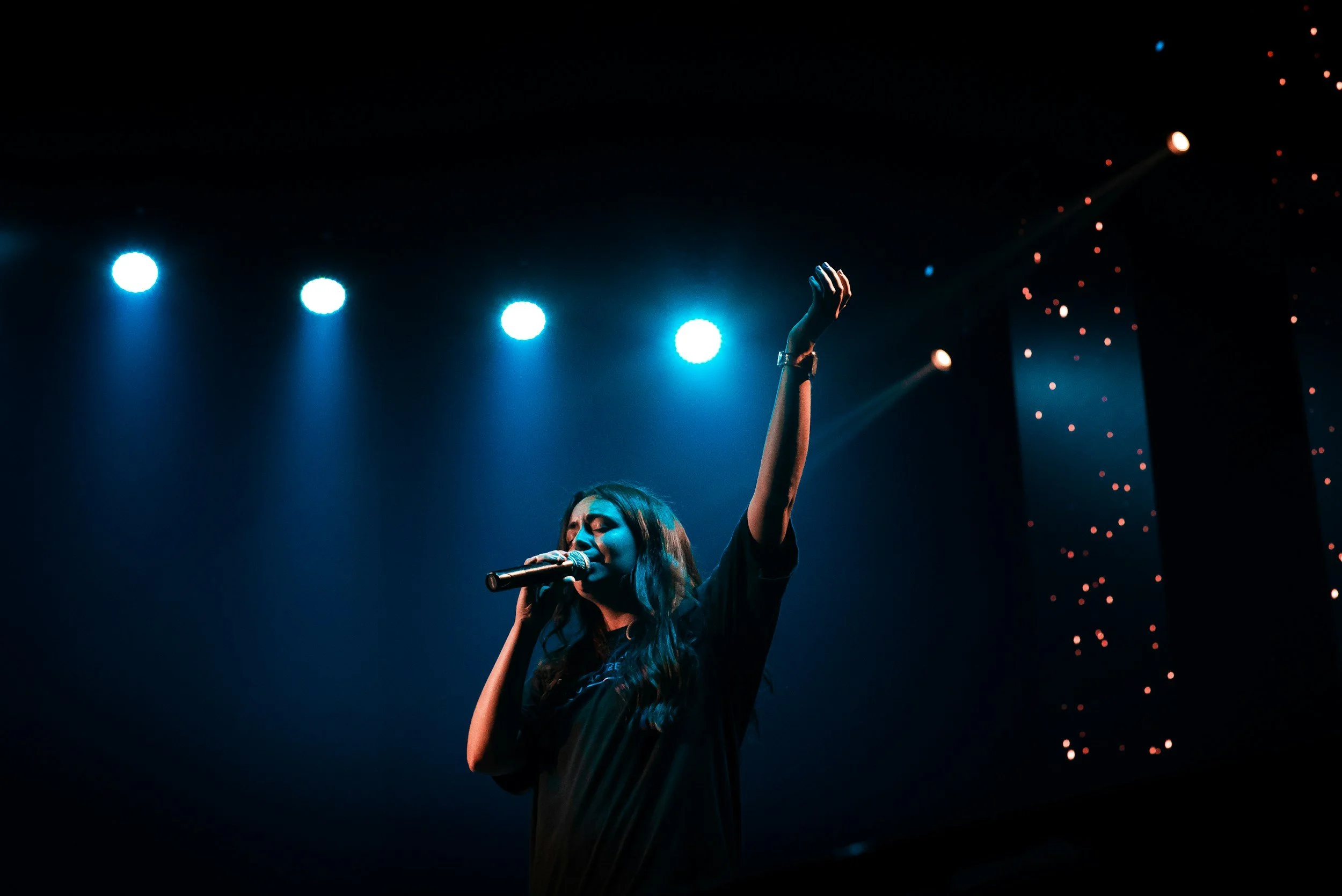 A female performer singing on stage with a microphone, under blue stage lights, and her arm raised.