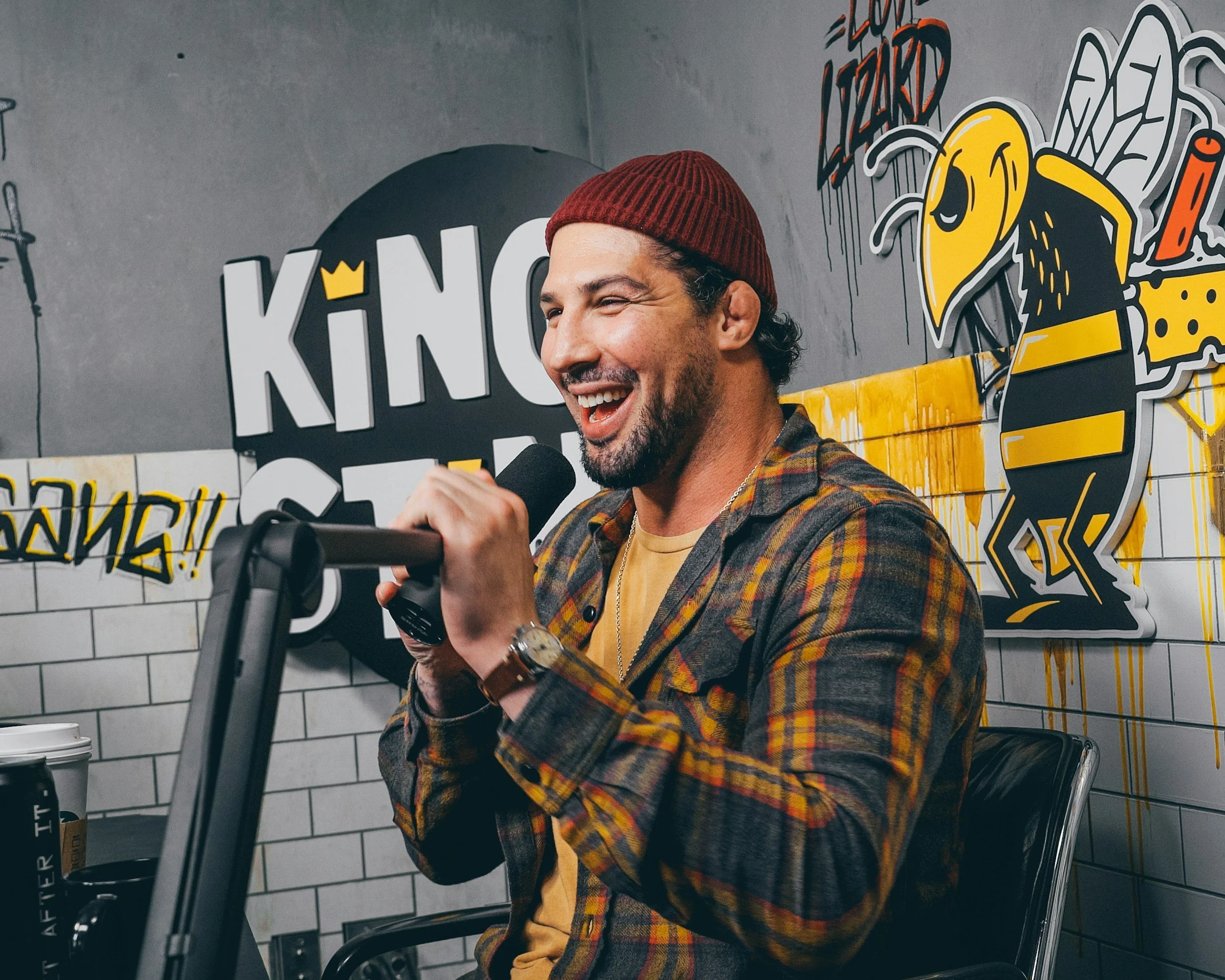 Man with a beard and a red beanie holding a microphone while smiling in a podcast studio with urban graffiti-style art and logo.