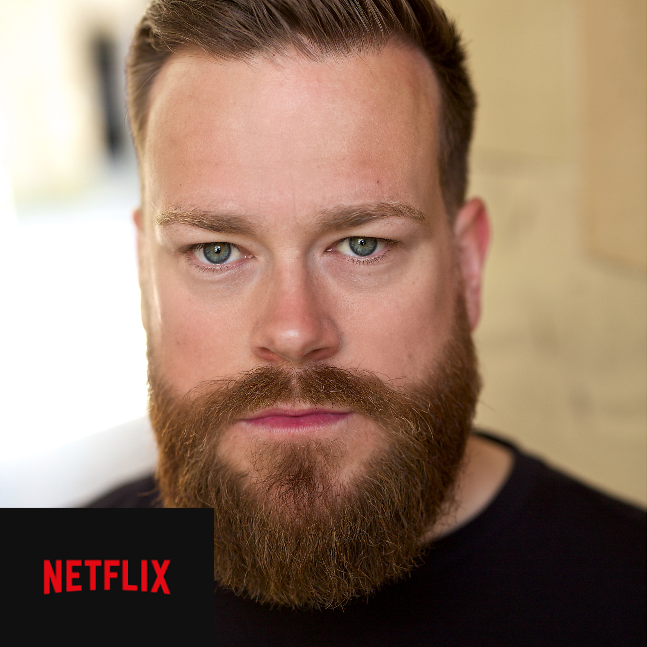 Close-up of a man with blue eyes, brown hair, and a full beard, looking directly at the camera, with a black shirt and a Netflix logo in the bottom left corner.