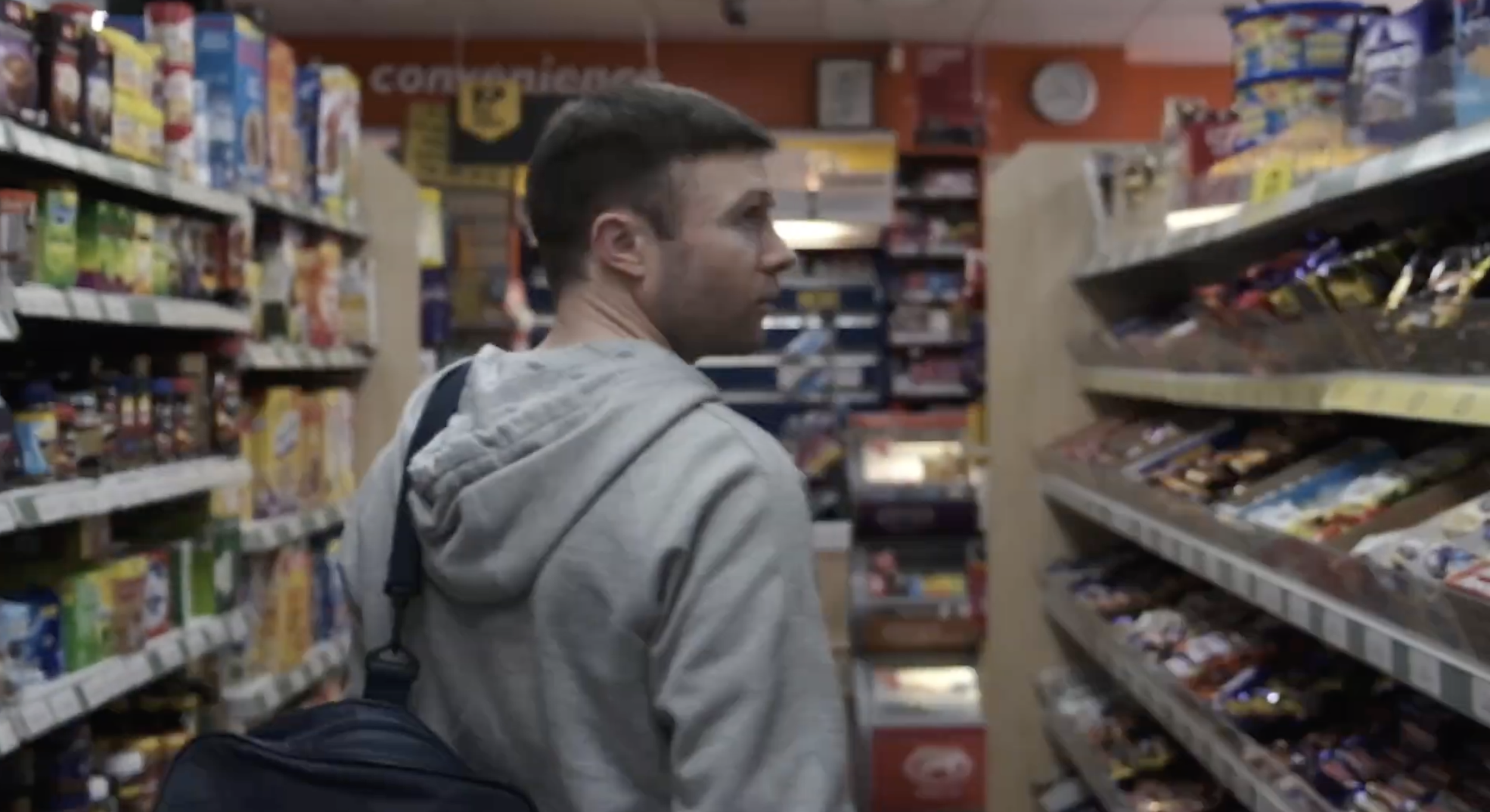 A man with short dark hair wearing a grey hoodie and a backpack shopping for snacks in a grocery store aisle.