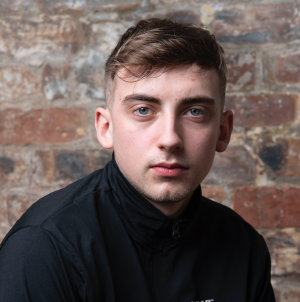 Portrait of a young man with short hair, wearing a black shirt, posing with arms crossed against a light background.