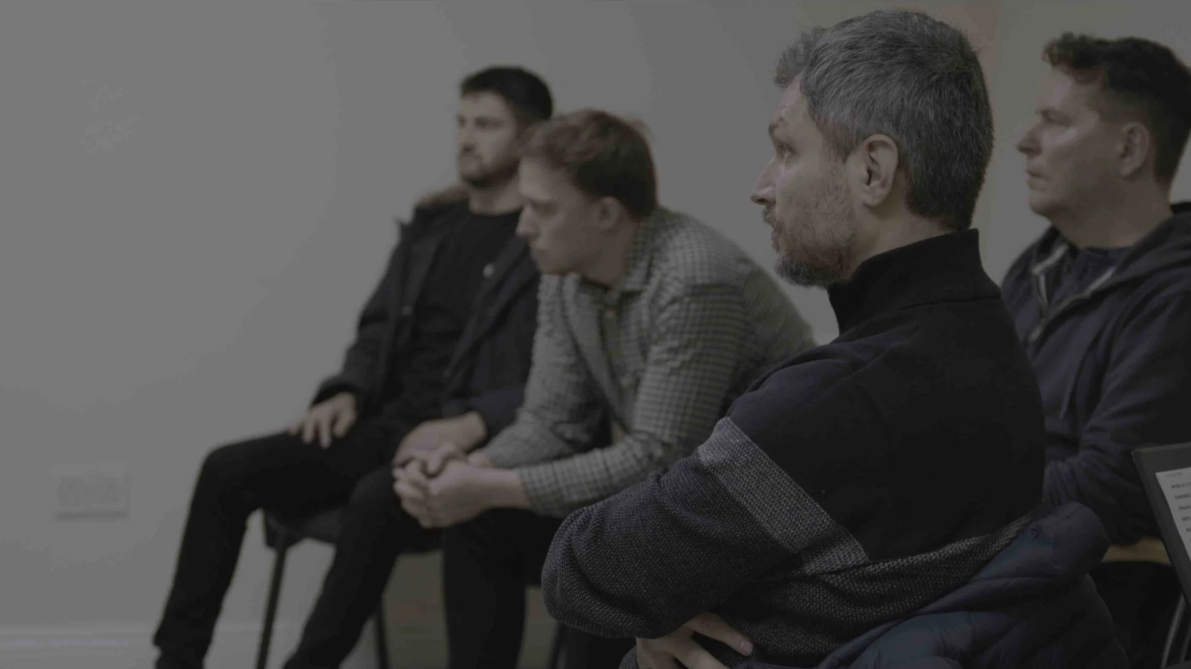 Five men seated in a row attending a meeting or presentation, all looking to the right, with neutral to focused expressions.