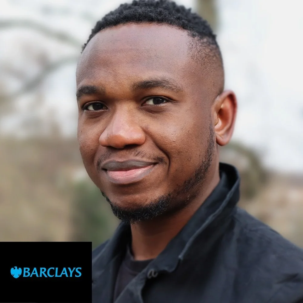 A young man with short curly hair and a slight beard, smiling at the camera with an outdoor background.