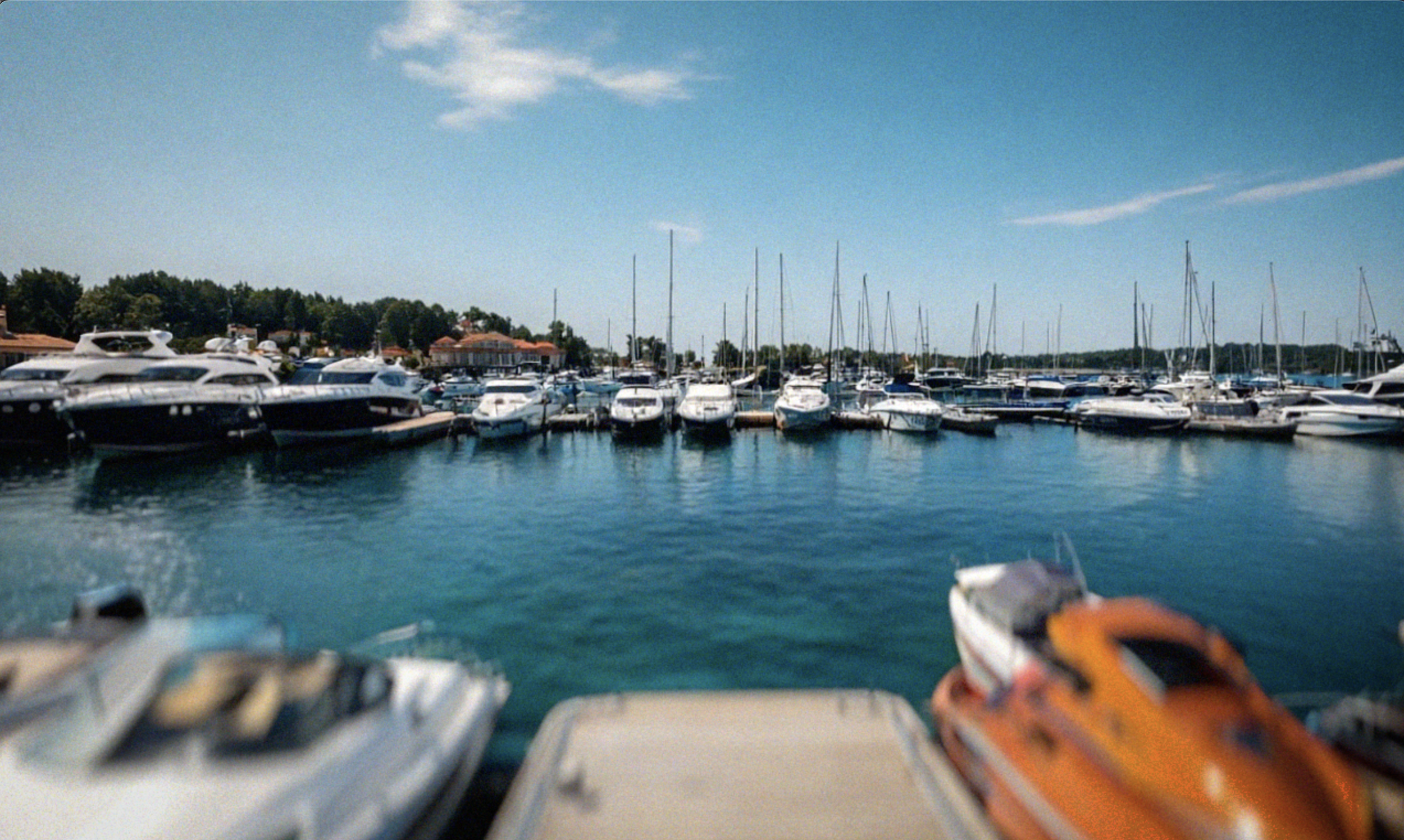 View of a marina with numerous boats docked, calm water, blue sky with a few clouds, and some trees and buildings in the background.