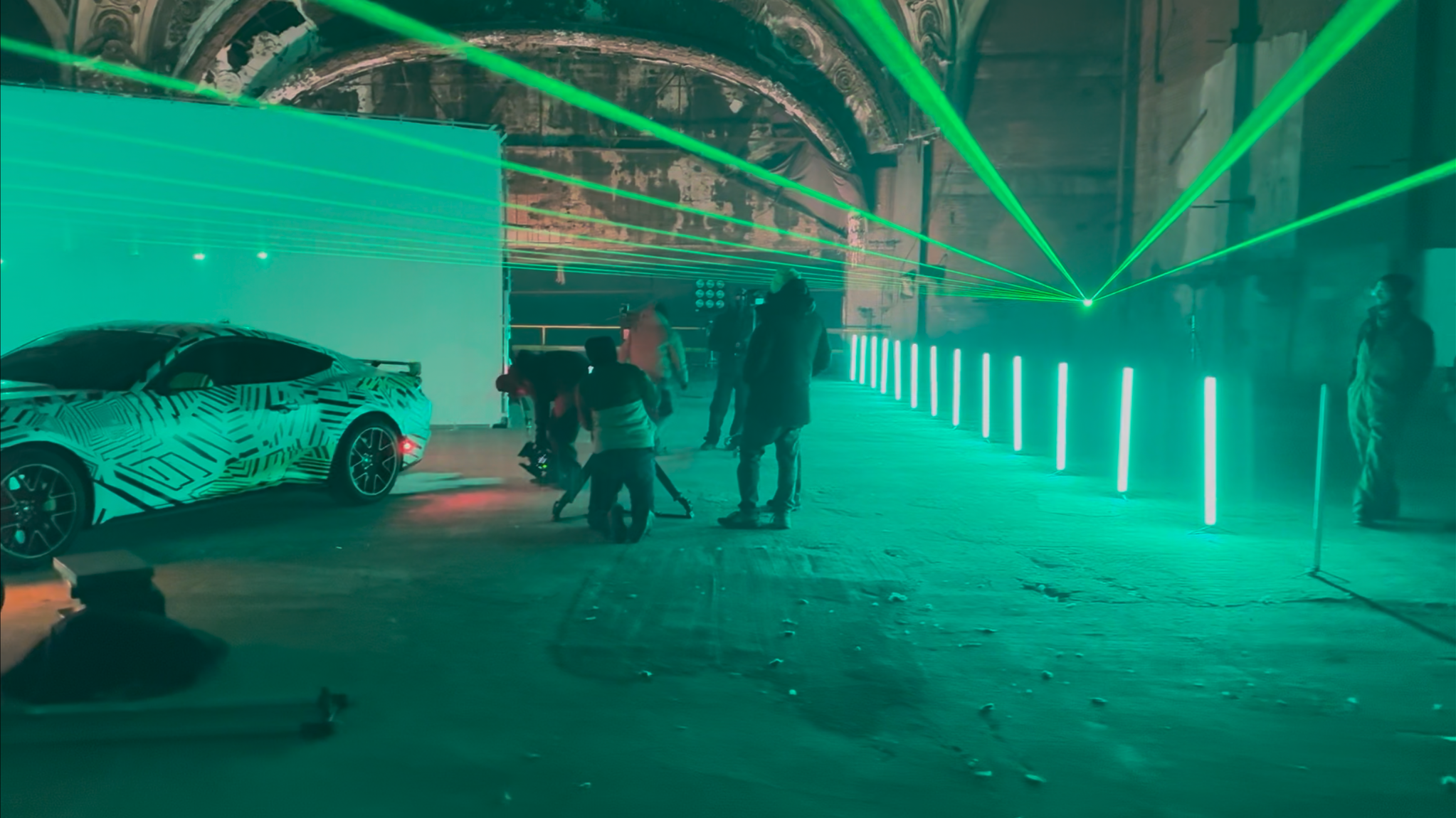 A car with a camouflage pattern is on display in a dimly lit industrial space with vibrant green laser lights and LED strip lights creating a futuristic atmosphere. Several people are gathered around, including some filming or photographing the scene