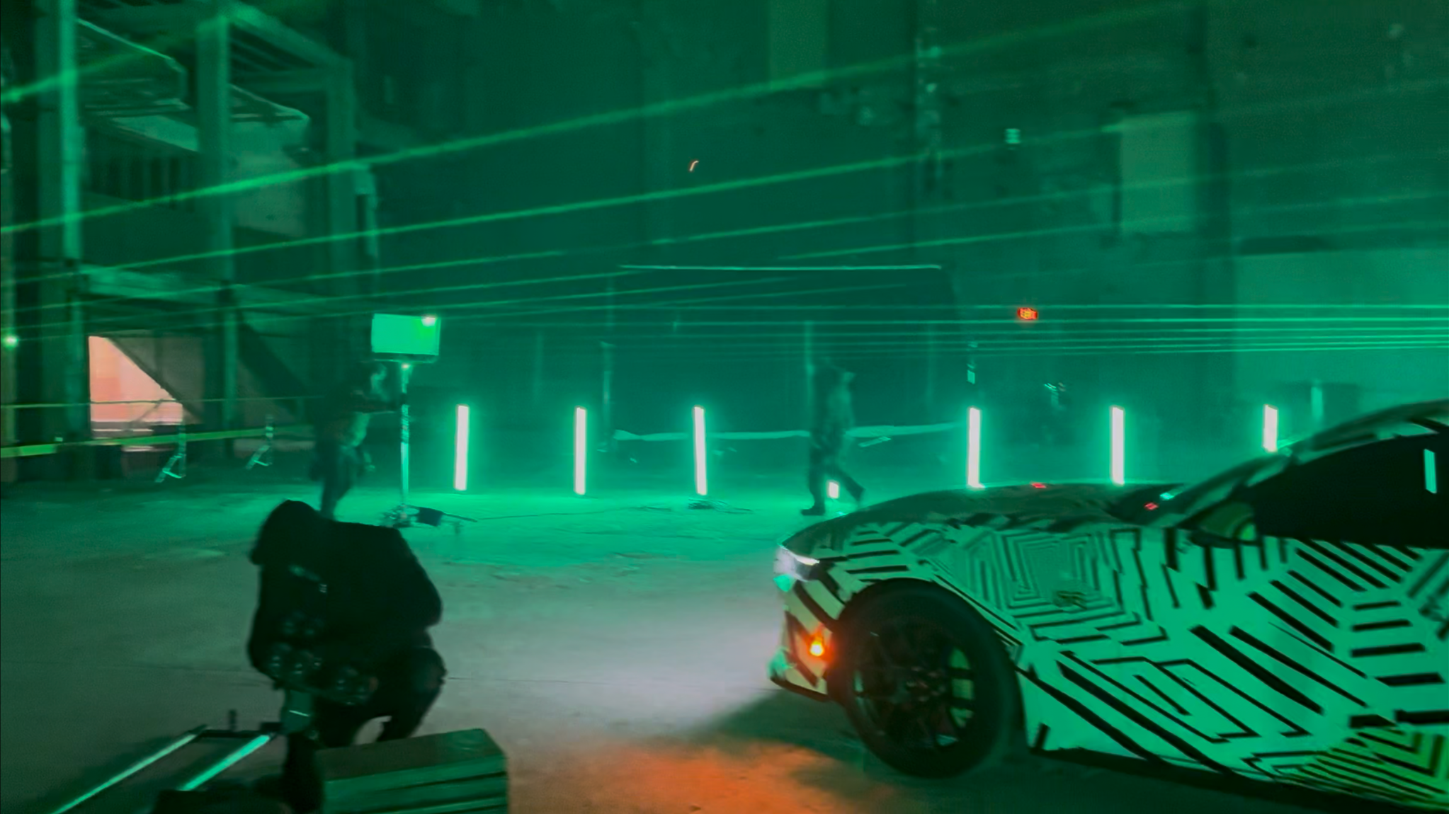 A photo taken at night showing a parking lot with green ambient lighting, a person crouching near the ground, a partially visible camouflaged vehicle in the foreground, and a few people walking among green vertical light poles.