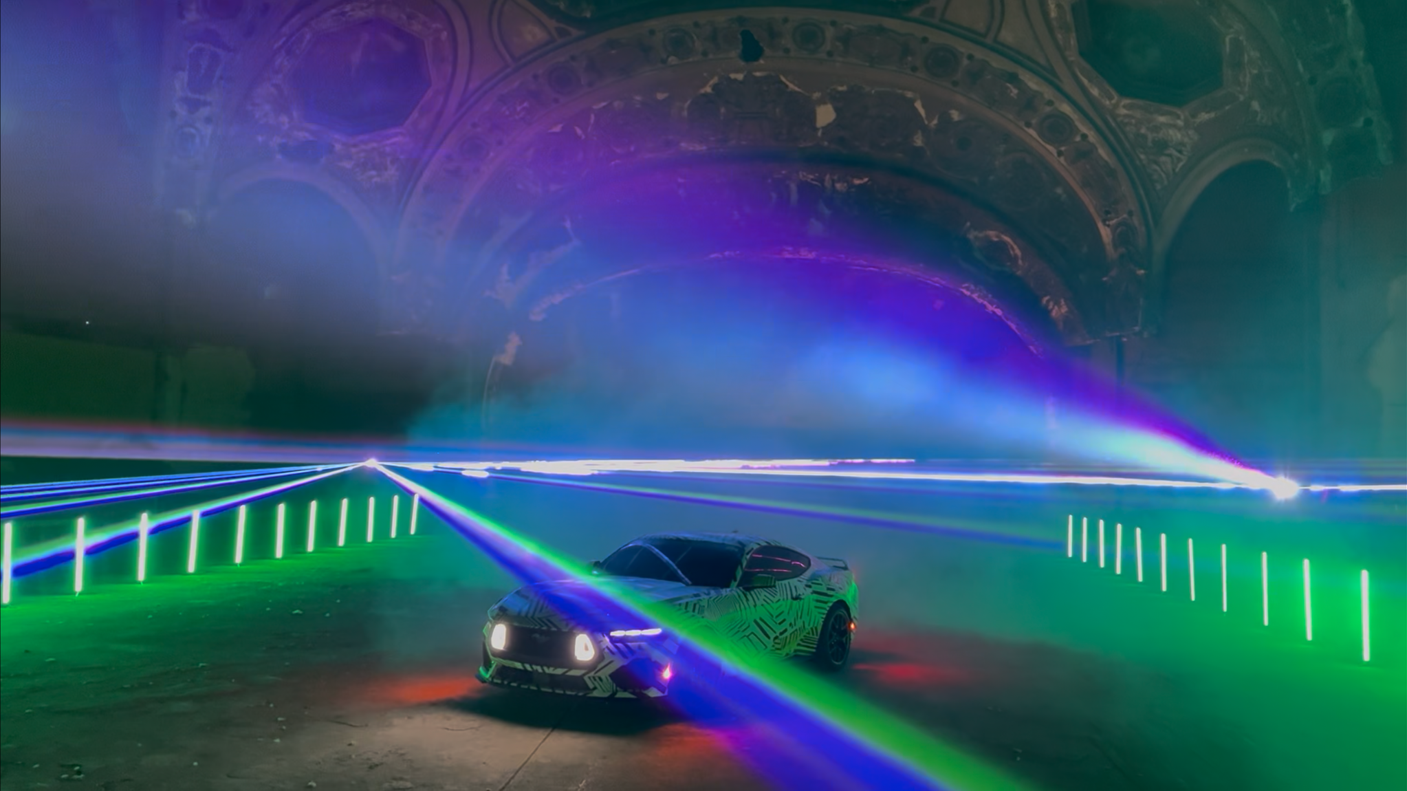 A futuristic sports car with a camouflage pattern under colorful laser lights inside a large industrial space or warehouse, with an arched ceiling visible in the background.
