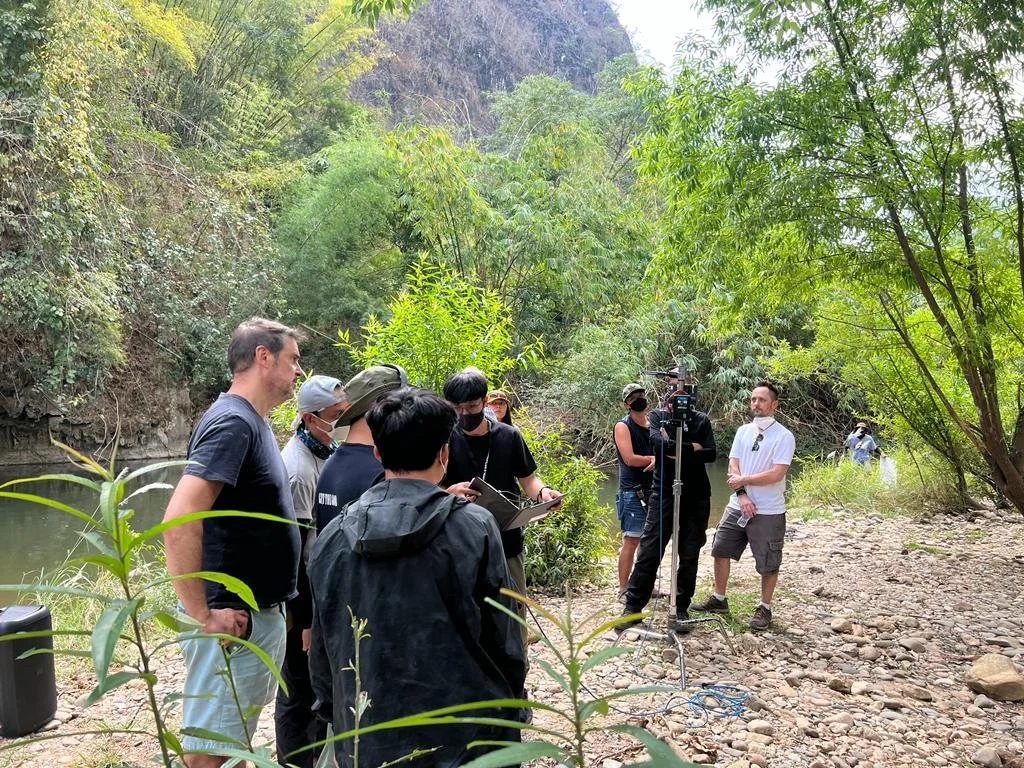Group of people standing outdoors in a forested area near a river, some wearing masks, looking at a camera and taking notes, with greenery and trees surrounding them.